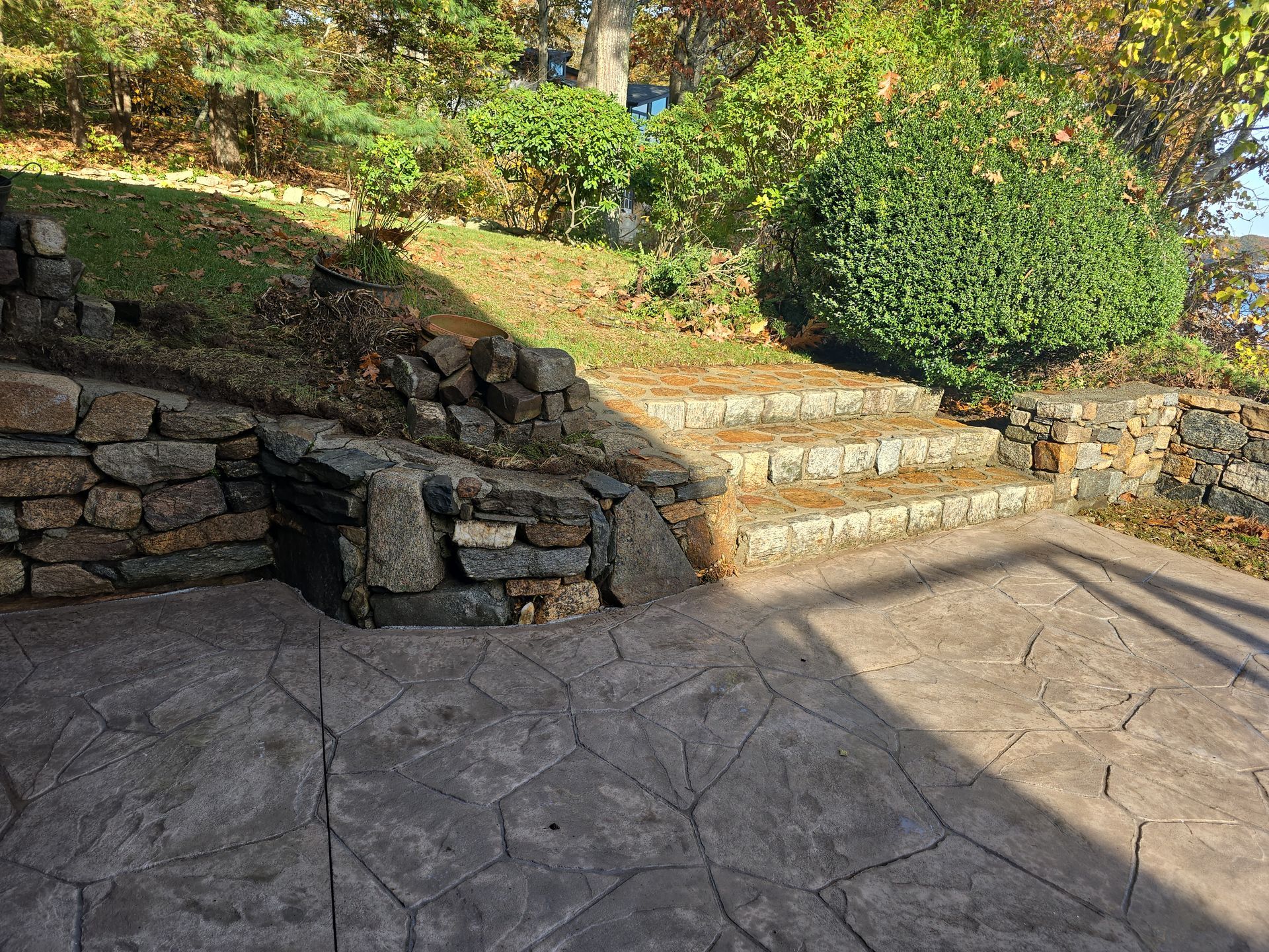 Stone patio with curved steps, shrubs, and a rock wall in a sunlit garden