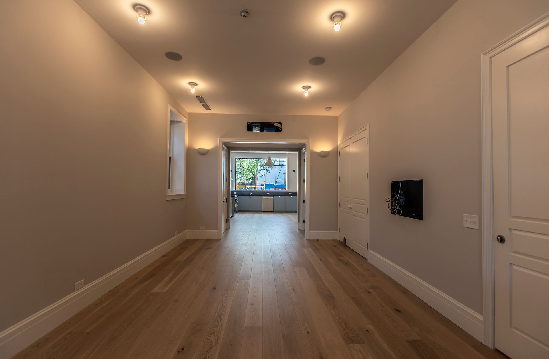 Long, empty hallway with wood floors, recessed lights, and a bright windowed room at the end.