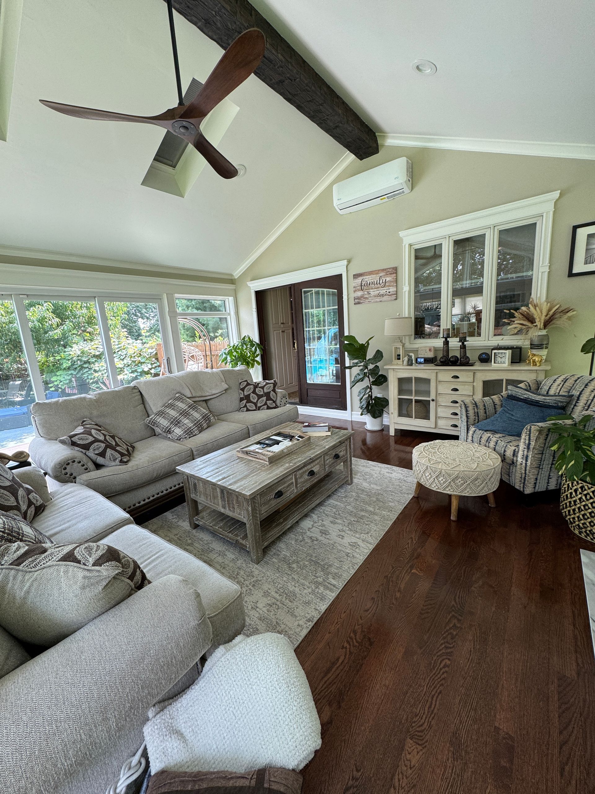 Bright sunroom living room with gray sectional, woven coffee table, dark wood floor, and open glass doors
