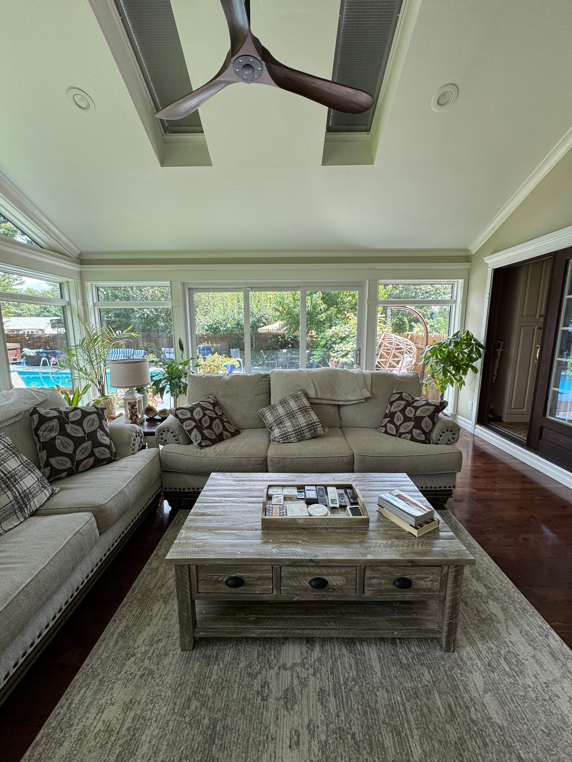 Bright living room with beige sectional sofa, wooden coffee table, large windows, and ceiling fan