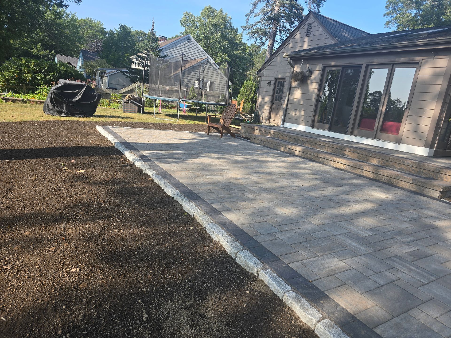 Paved patio beside a raised garden bed and house, with a chair and sunlit backyard view.