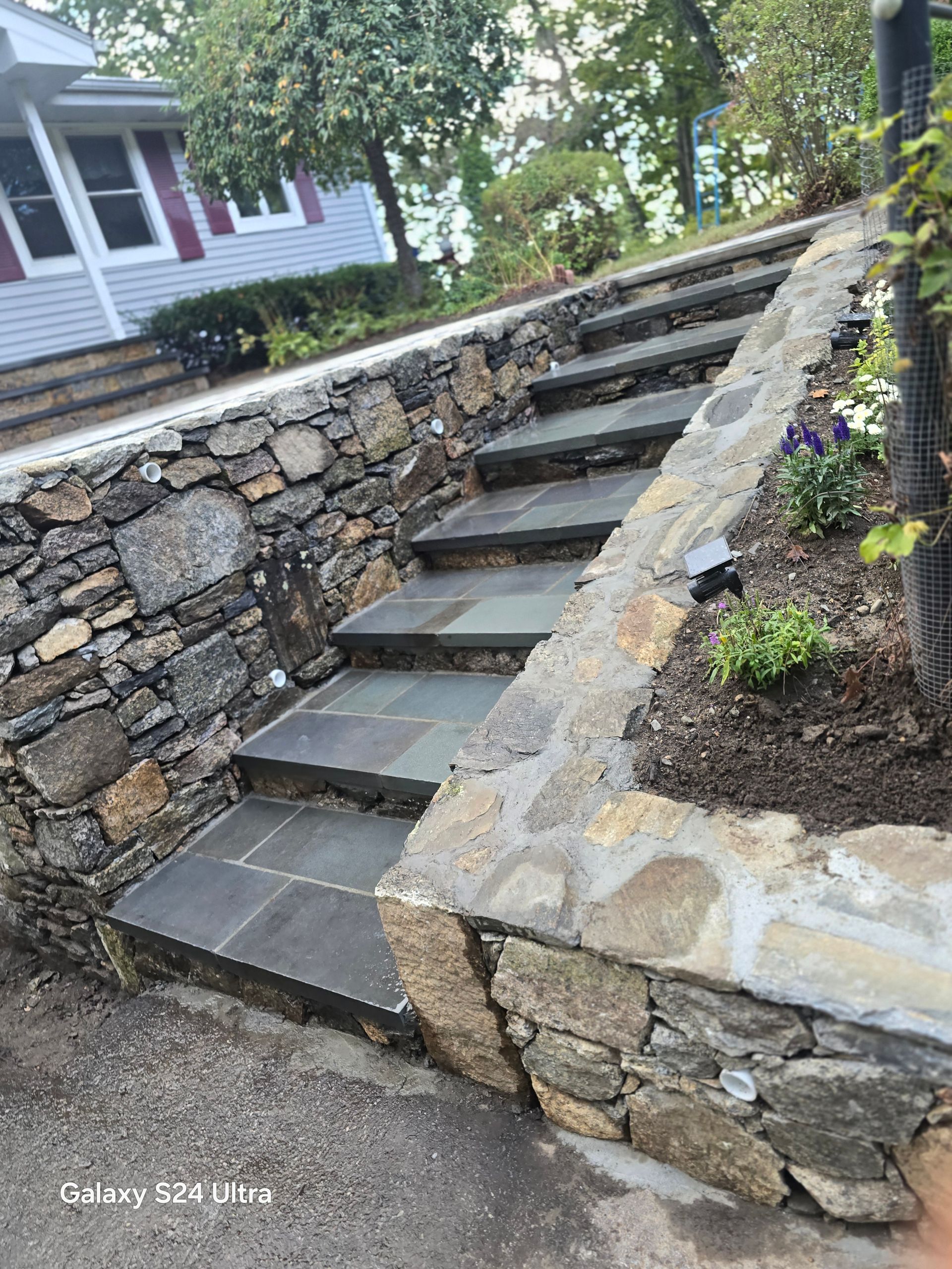 Stone outdoor stairs between retaining walls beside a house and garden bed