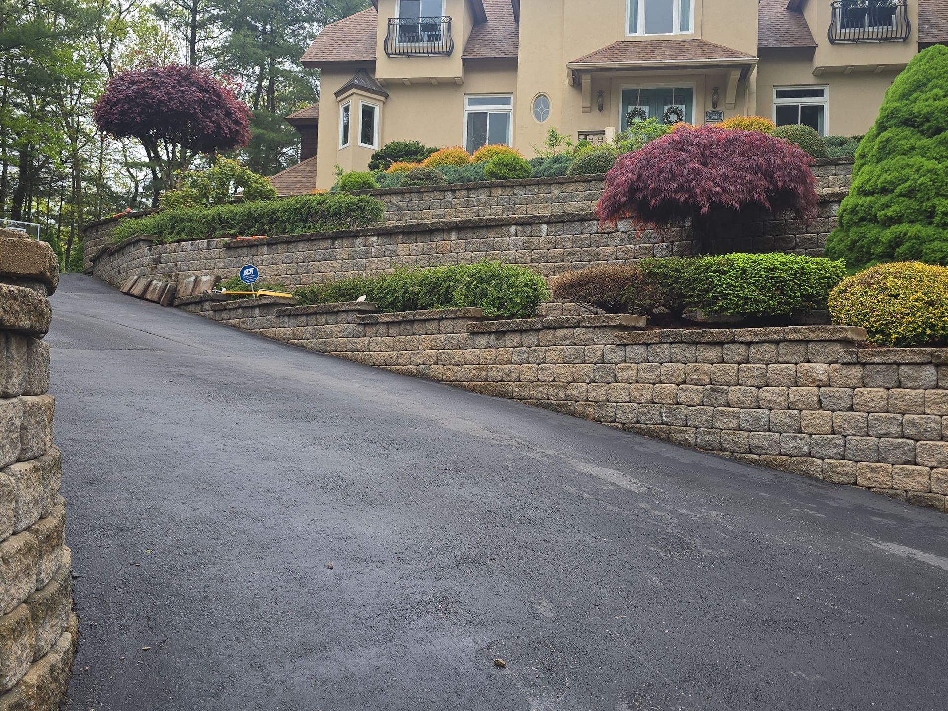 Curved driveway beside stone retaining walls and landscaped shrubs leading to a beige house