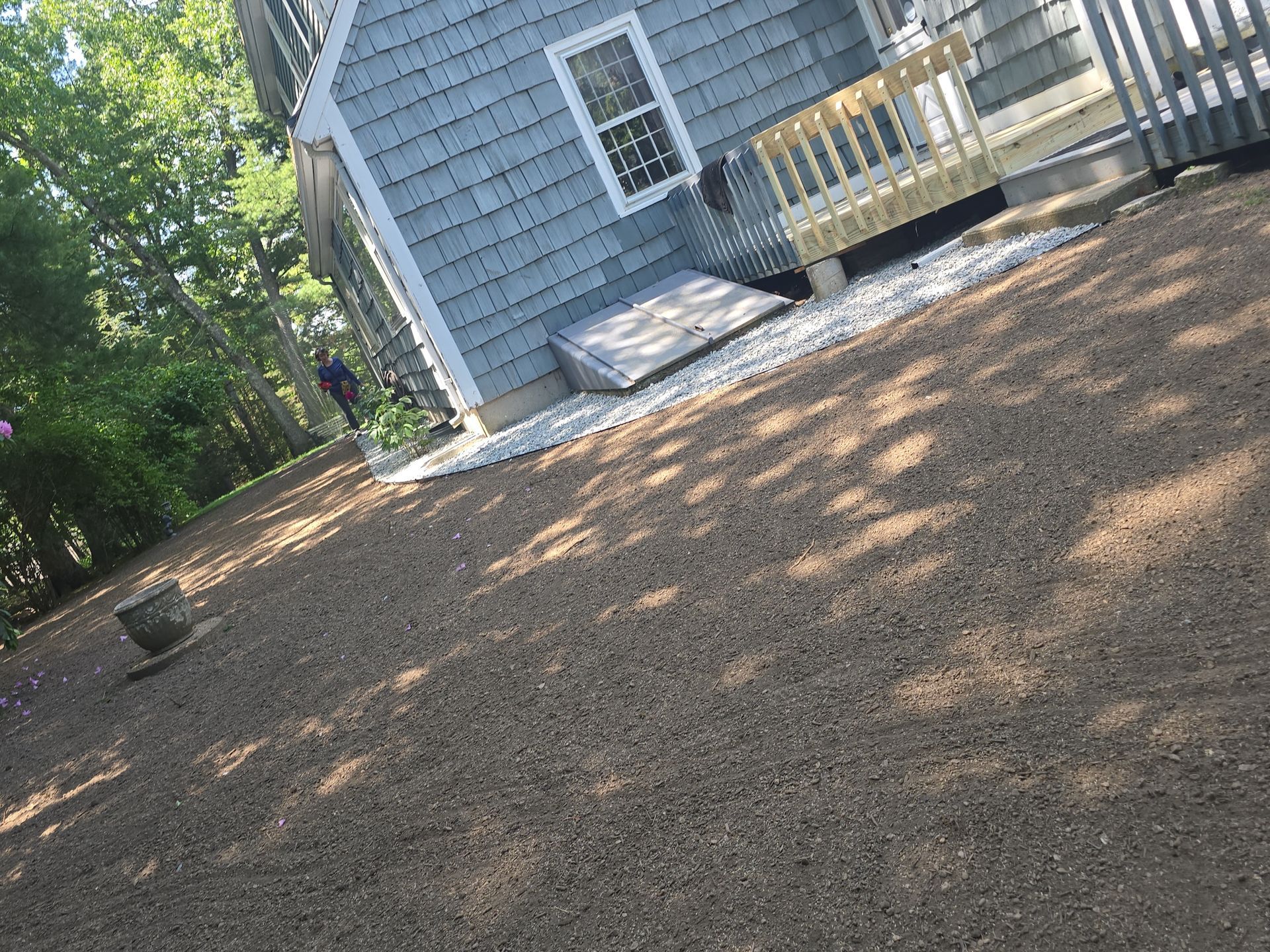 Tilted view of a gray house with a small porch and ramp beside a sunlit gravel yard