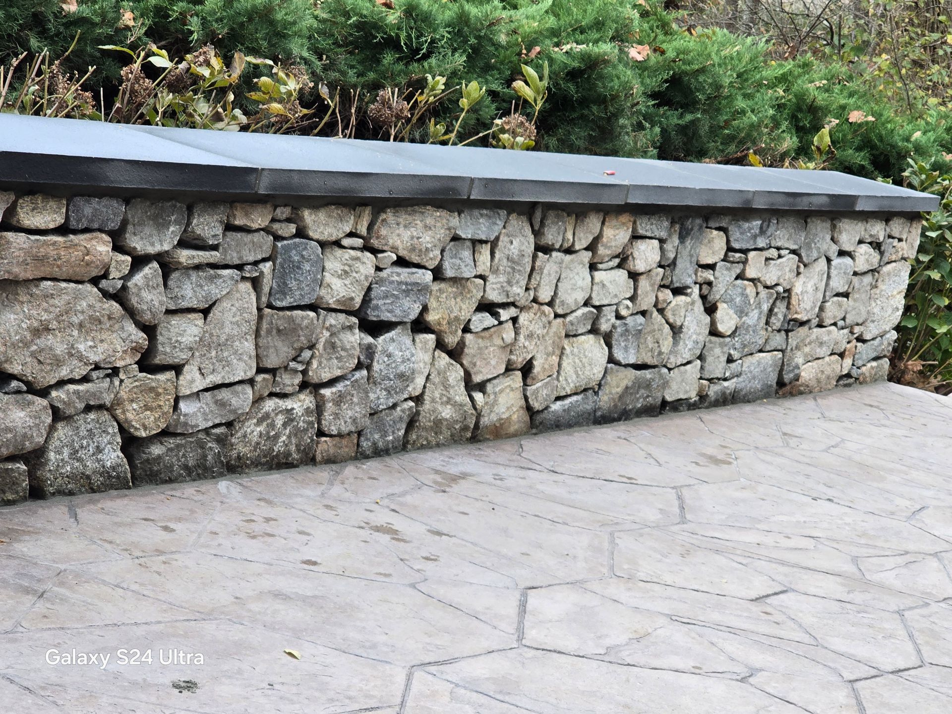 Stone retaining wall beside a light-colored paved walkway with greenery above it