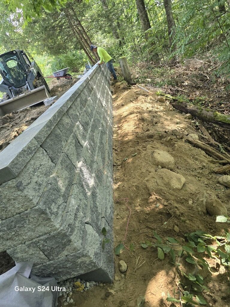 Person on a dirt path beside a stone wall, with an excavator in a wooded area.