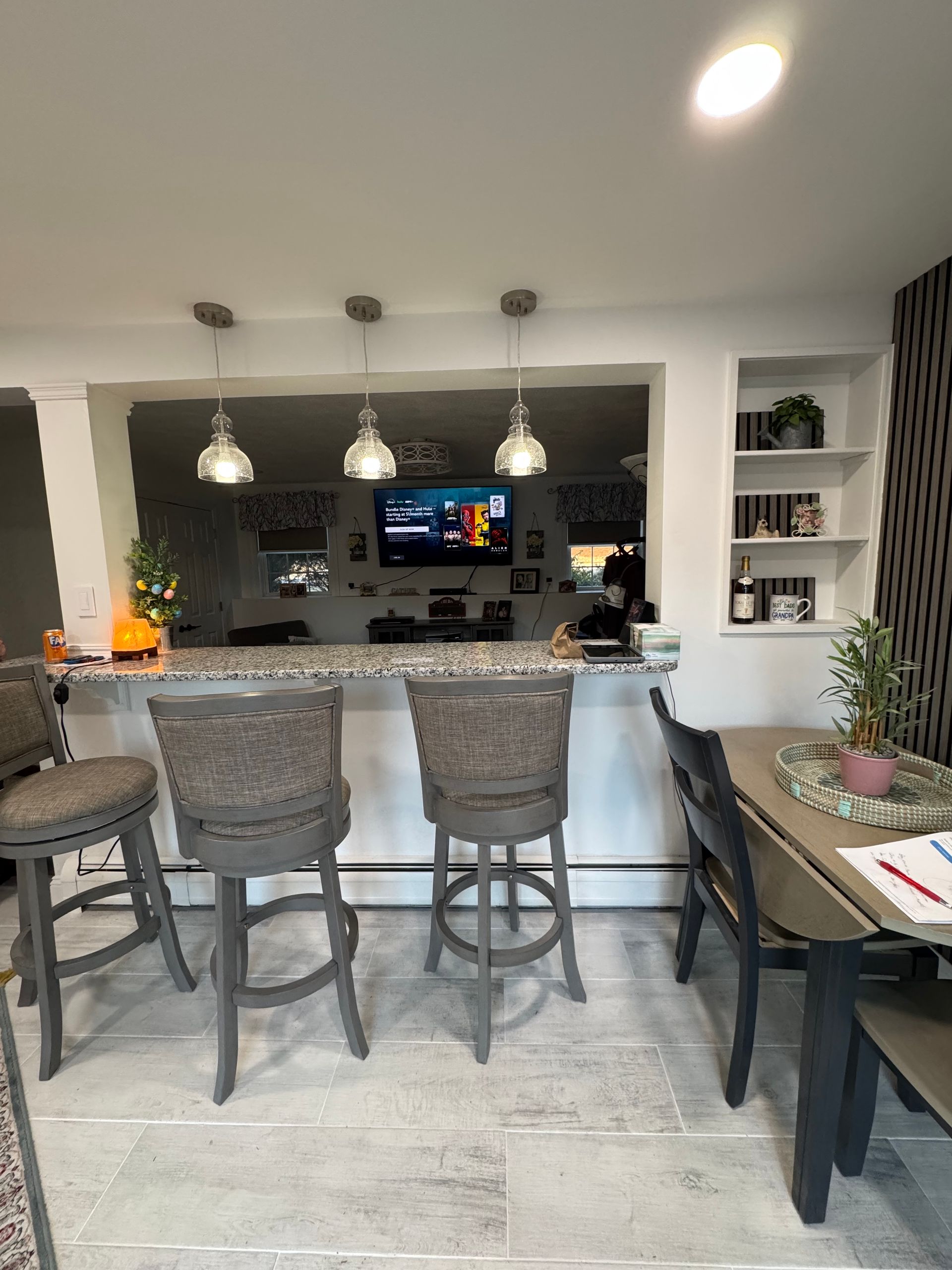 Modern kitchen with granite island, three woven bar stools, pendant lights, and a TV visible through a pass-through opening