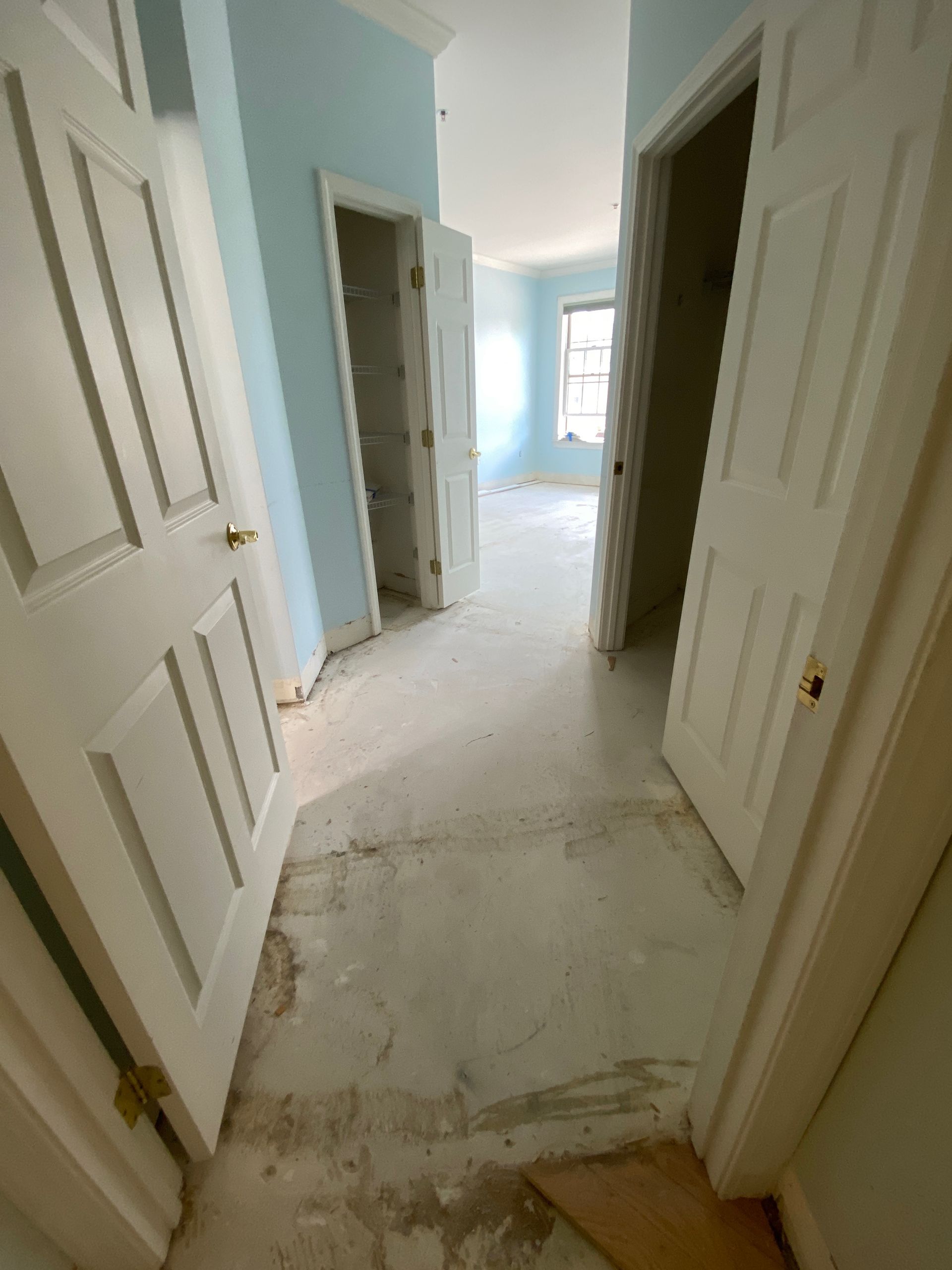 Narrow hallway with open doors and unfinished floors, leading to a bright room at the end.