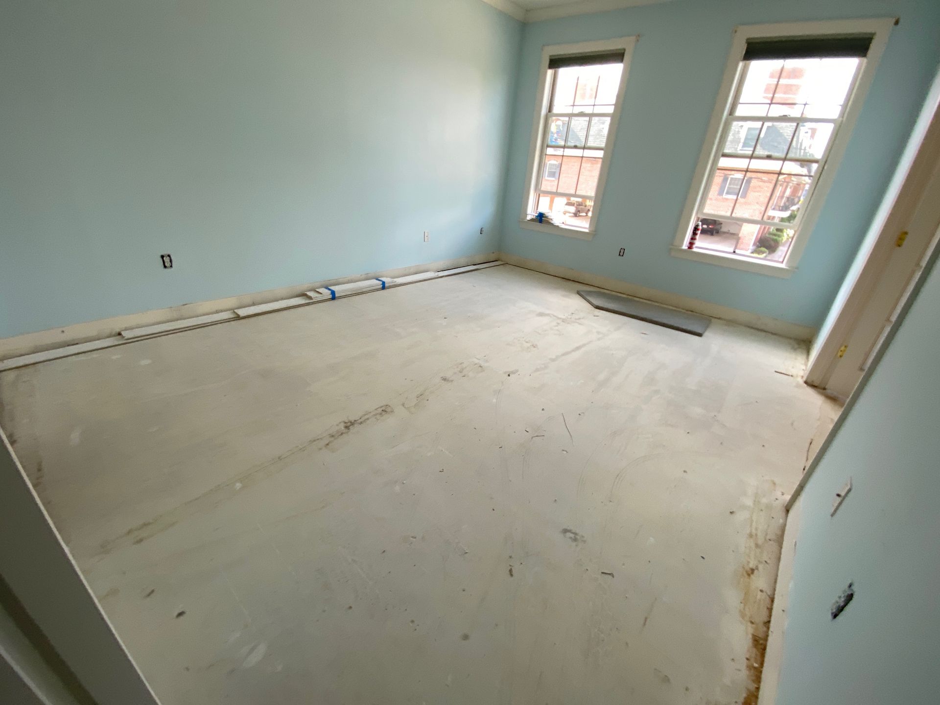 Empty light-blue room with unfinished floor and two windows letting in daylight