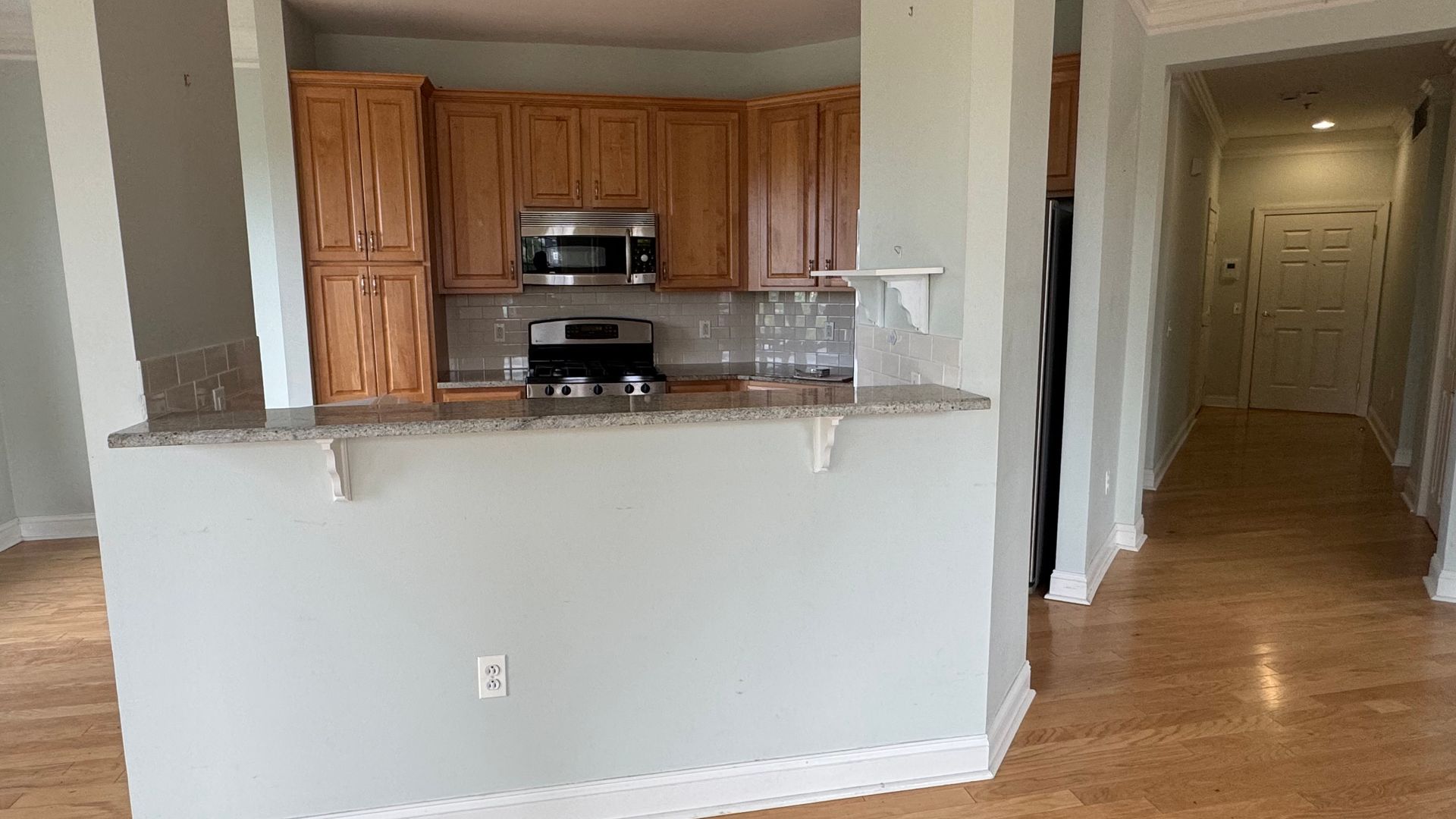 Empty kitchen pass-through with wood cabinets and stainless appliances, opening to a hallway with hardwood floors.