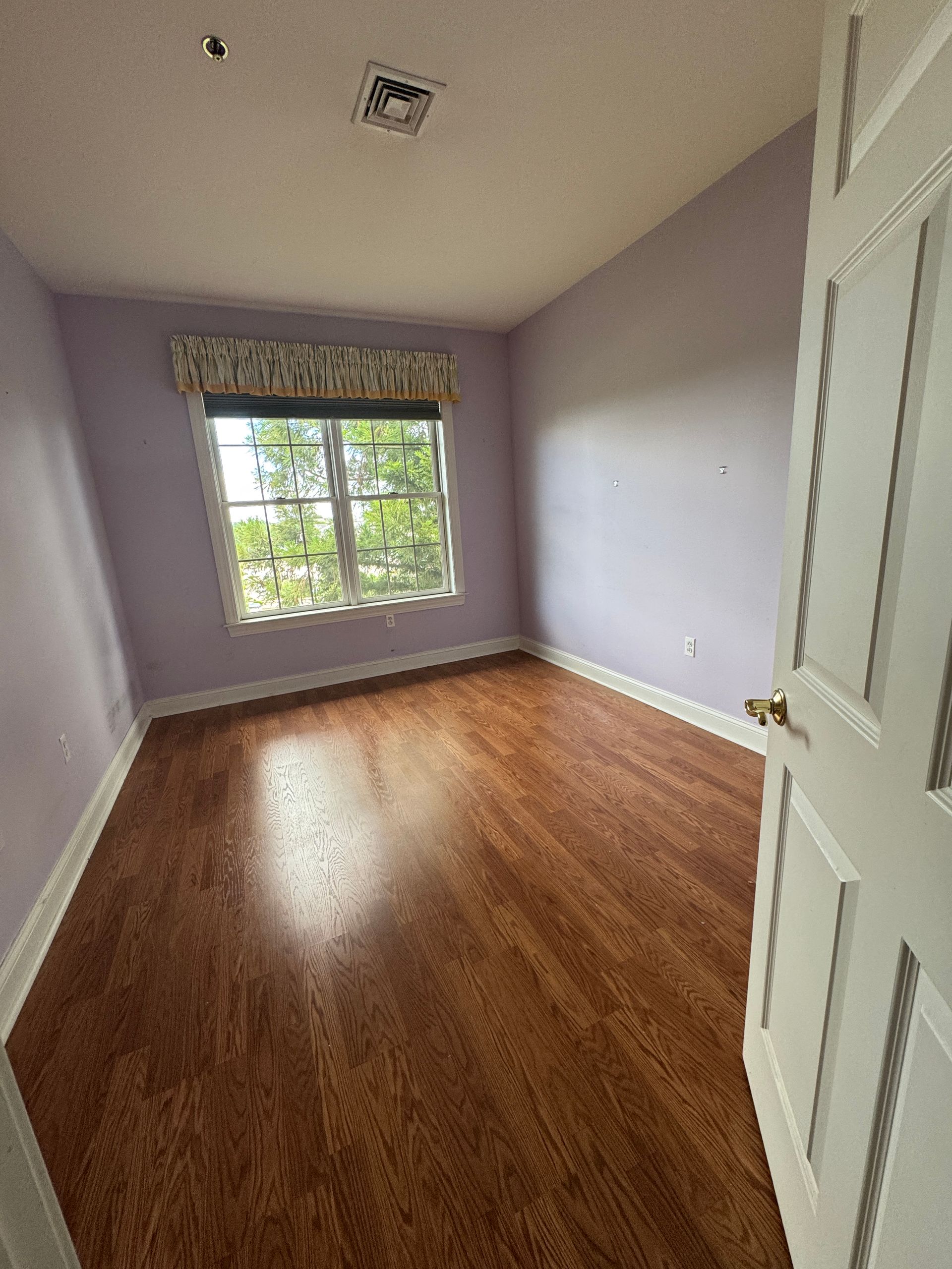 Empty lavender bedroom with wood floors, a window seat, and an open door.