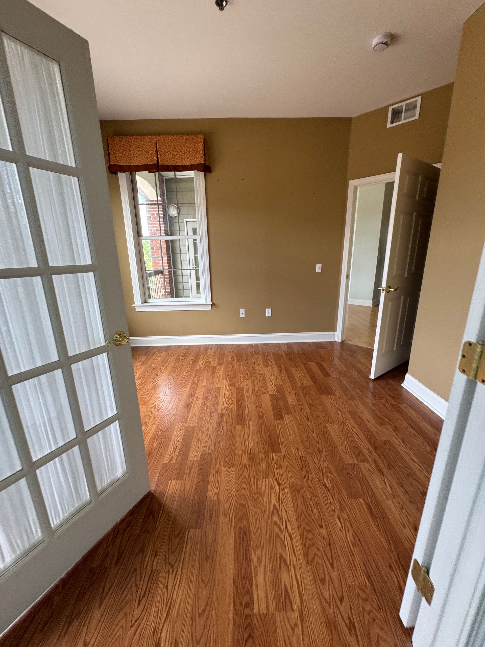 Empty beige room with wood floors, French doors, and a window with a valance.