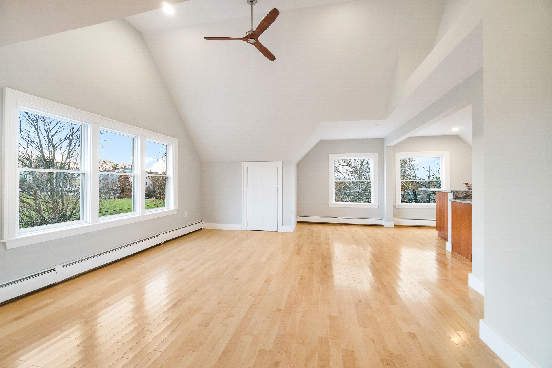 Bright, empty living room with hardwood floors, large windows, vaulted white ceiling, and ceiling fan.