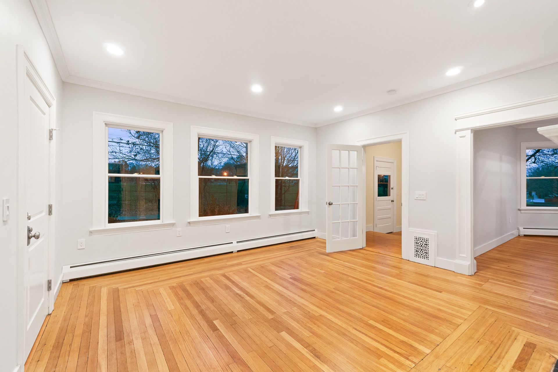 Empty bright living room with hardwood floors, white walls, and large windows at dusk