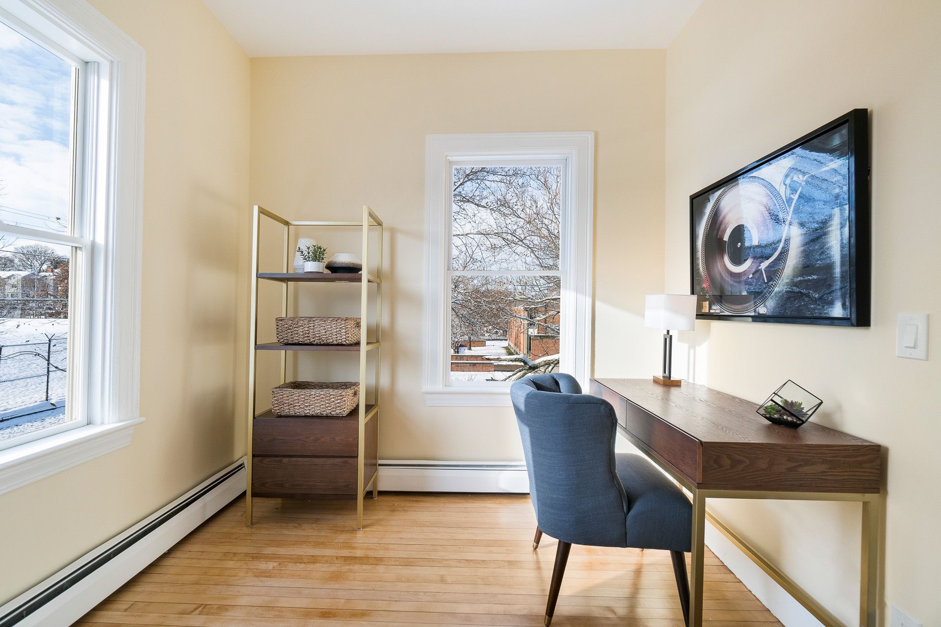 Bright home office with desk, blue chair, wall art, shelving, and two windows letting in daylight