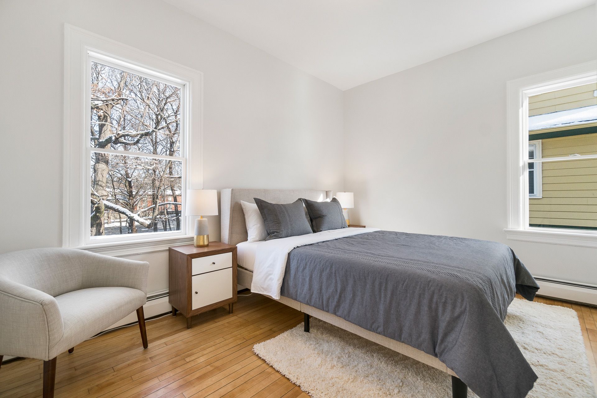 Bright minimalist bedroom with a bed, armchair, nightstand, and two windows letting in daylight.