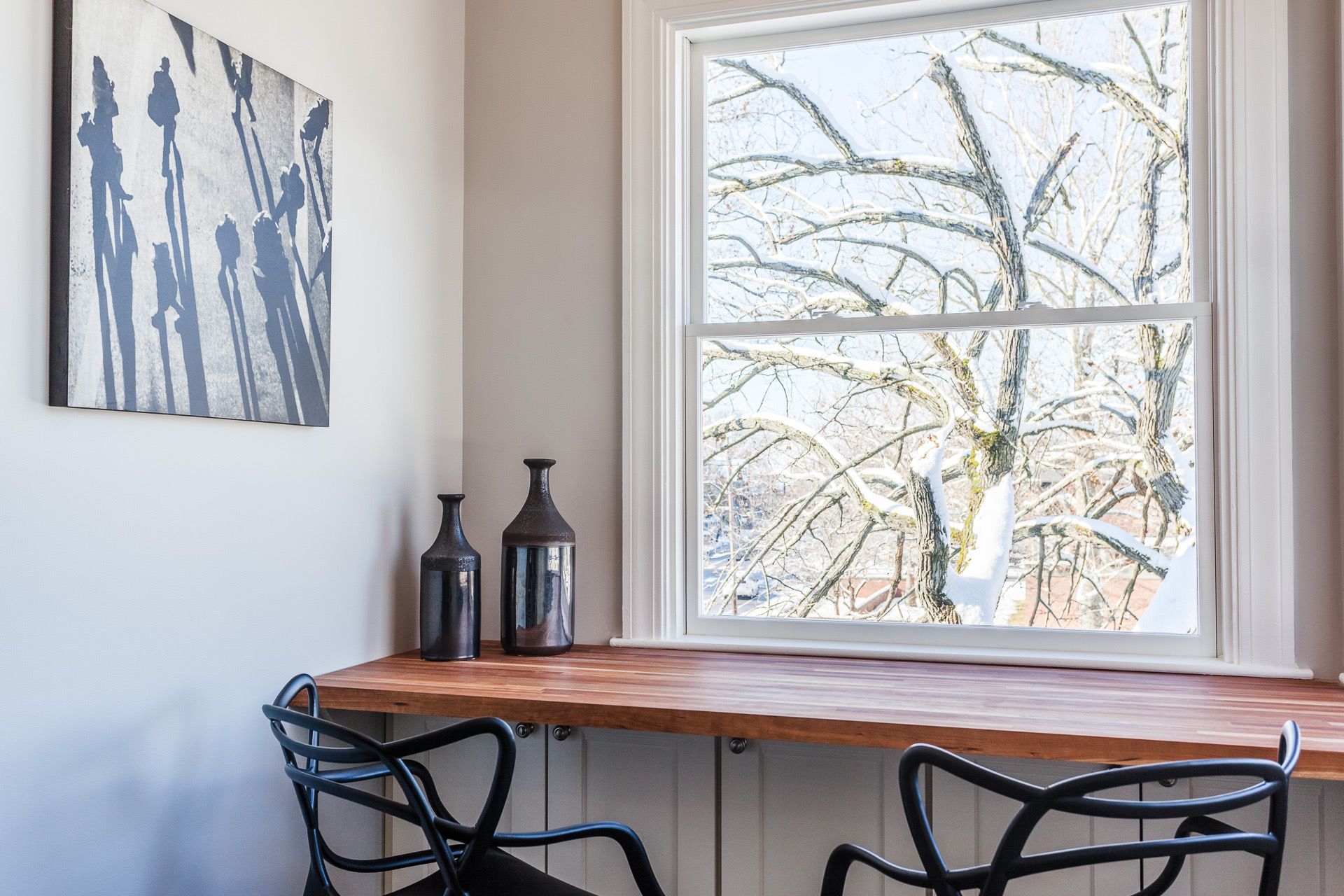 Bright home office nook with a wooden desk, black chairs, wall art, and a large window looking out to bare branches