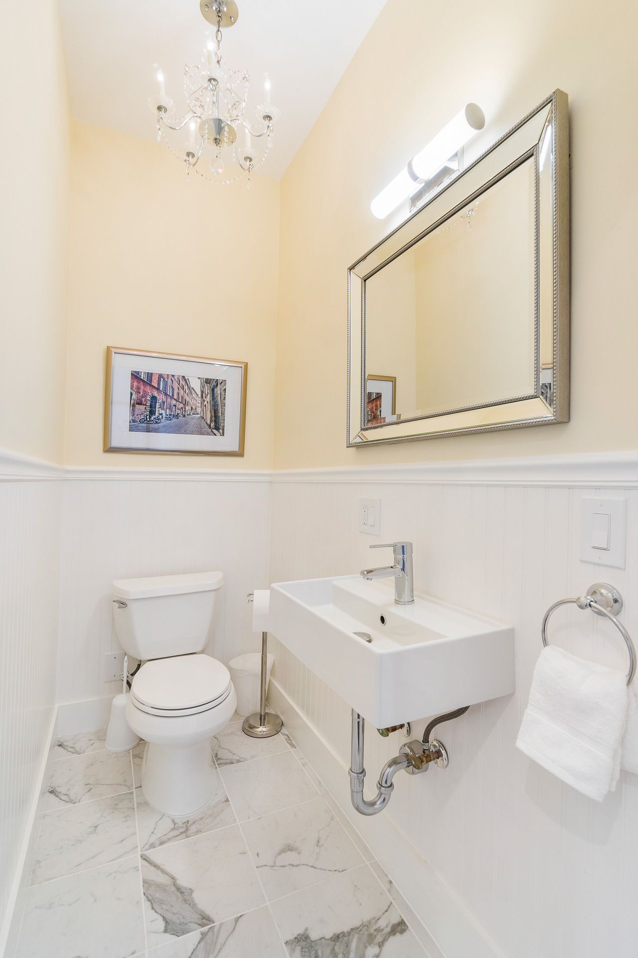 Elegant white bathroom with pedestal sink, toilet, large mirror, wall sconce, and chandelier