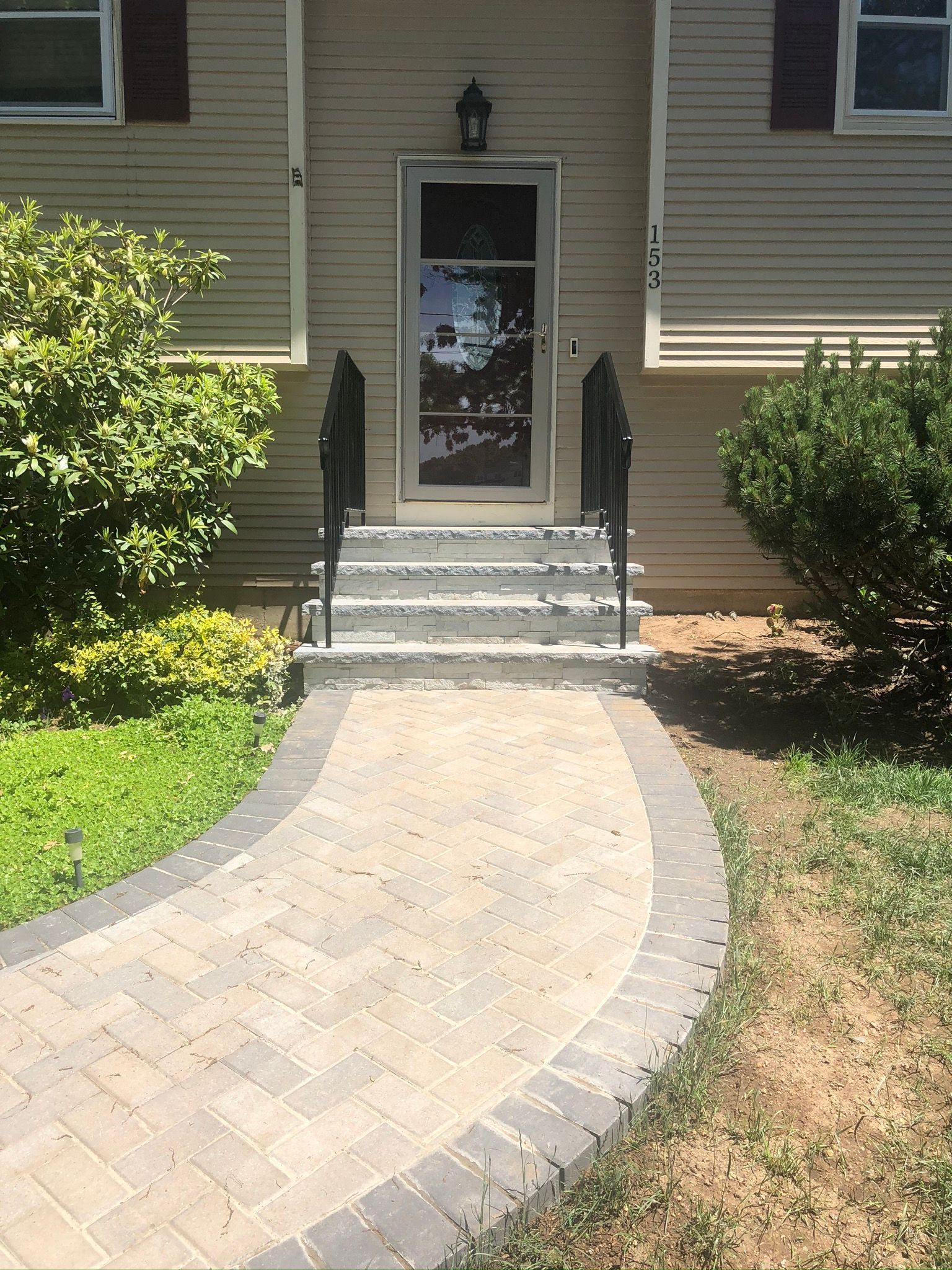 Front entrance with curved paver walkway, steps, black railings, and a glass door between shrubs.