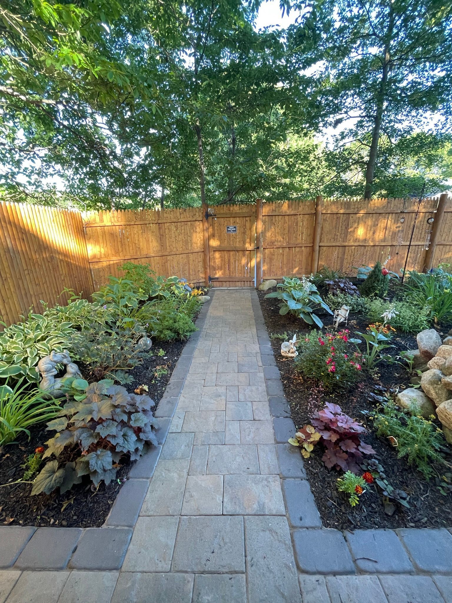 Stone garden path leading to a wooden gate, bordered by lush plants and flowers in a backyard.