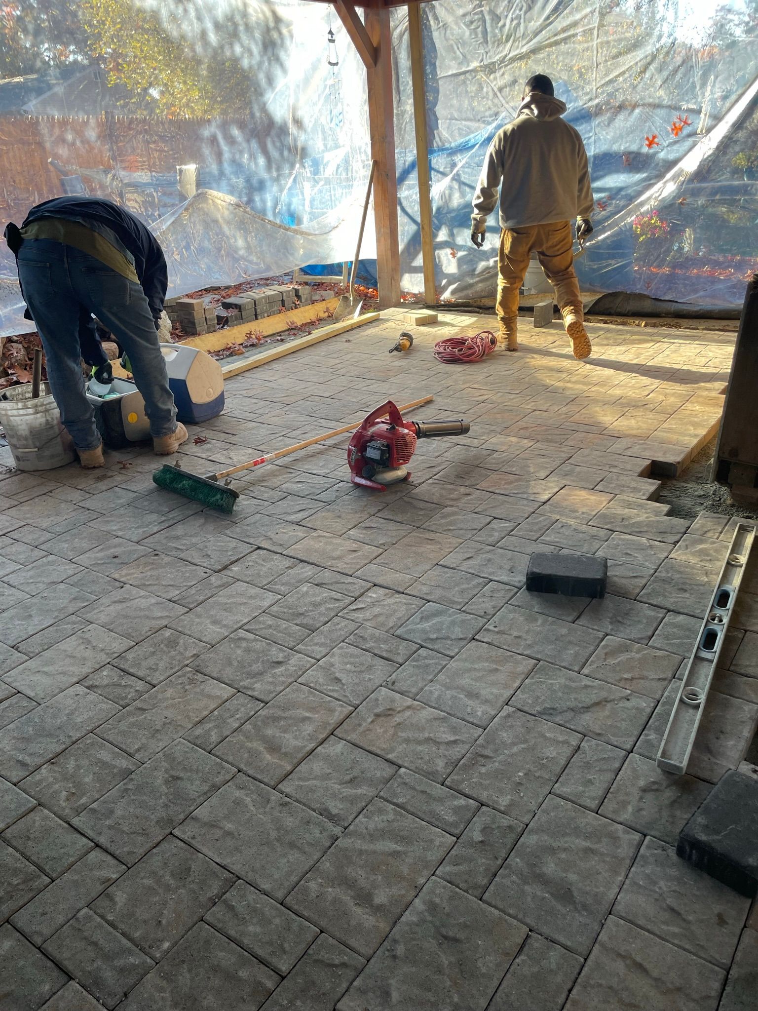 Workers installing paver stones on a patio, with tools and debris scattered across the ground.