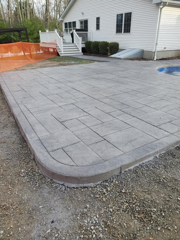 Freshly paved gray patio beside a white house with steps, orange fencing, and gravel border