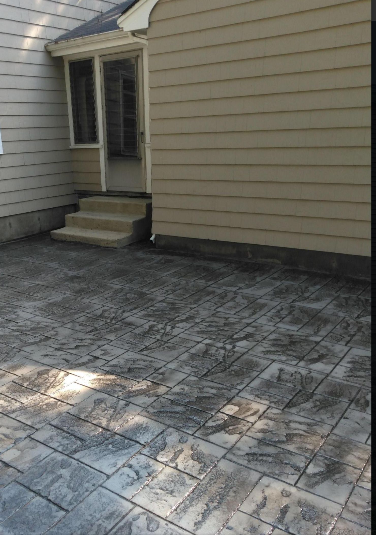 Small concrete patio beside a beige house with a back door and steps, lit by sunlight.