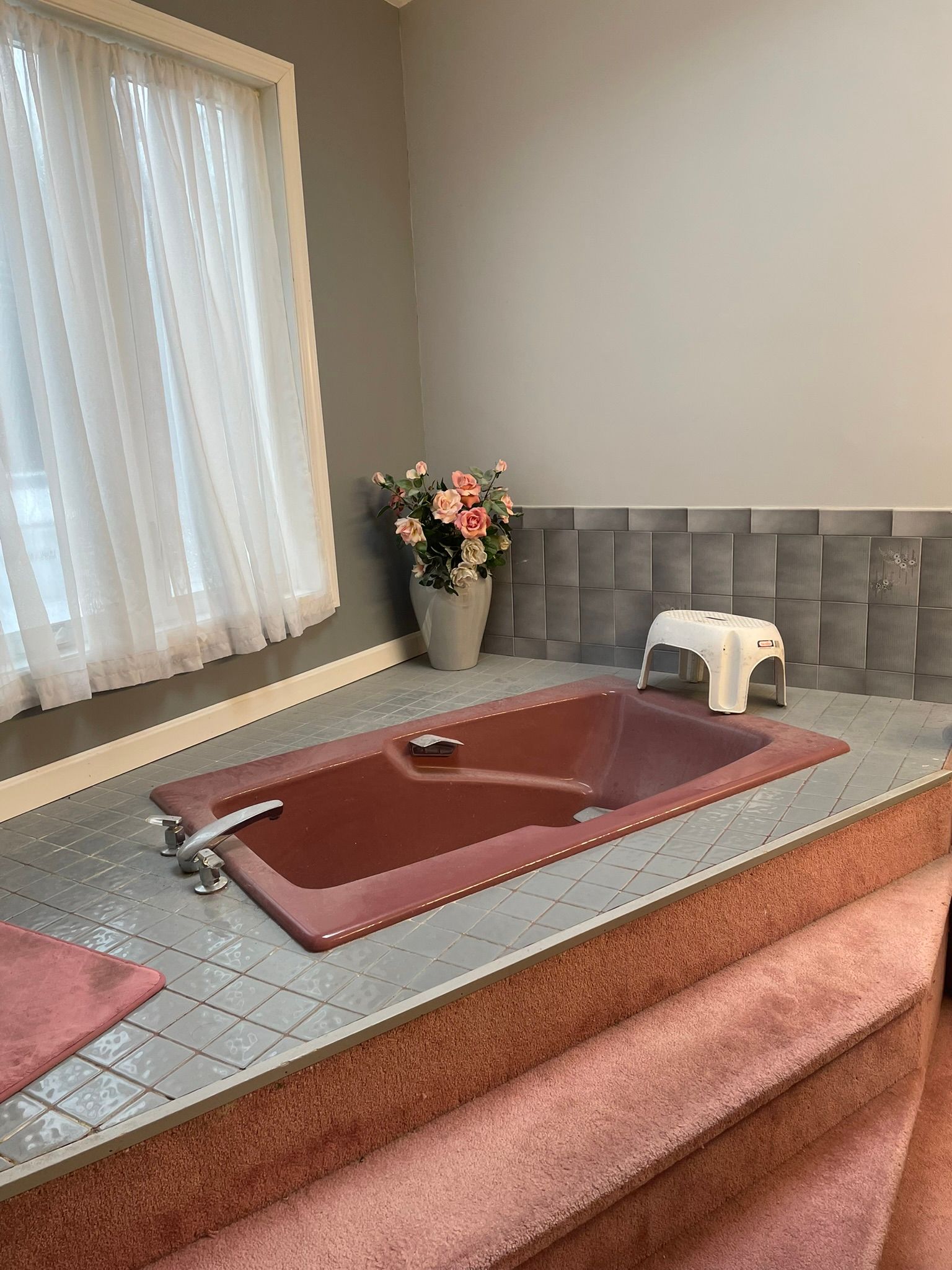 Pink bathroom sink with chrome faucet, gray tiled counter, window with sheer curtains, and flowers in the corner