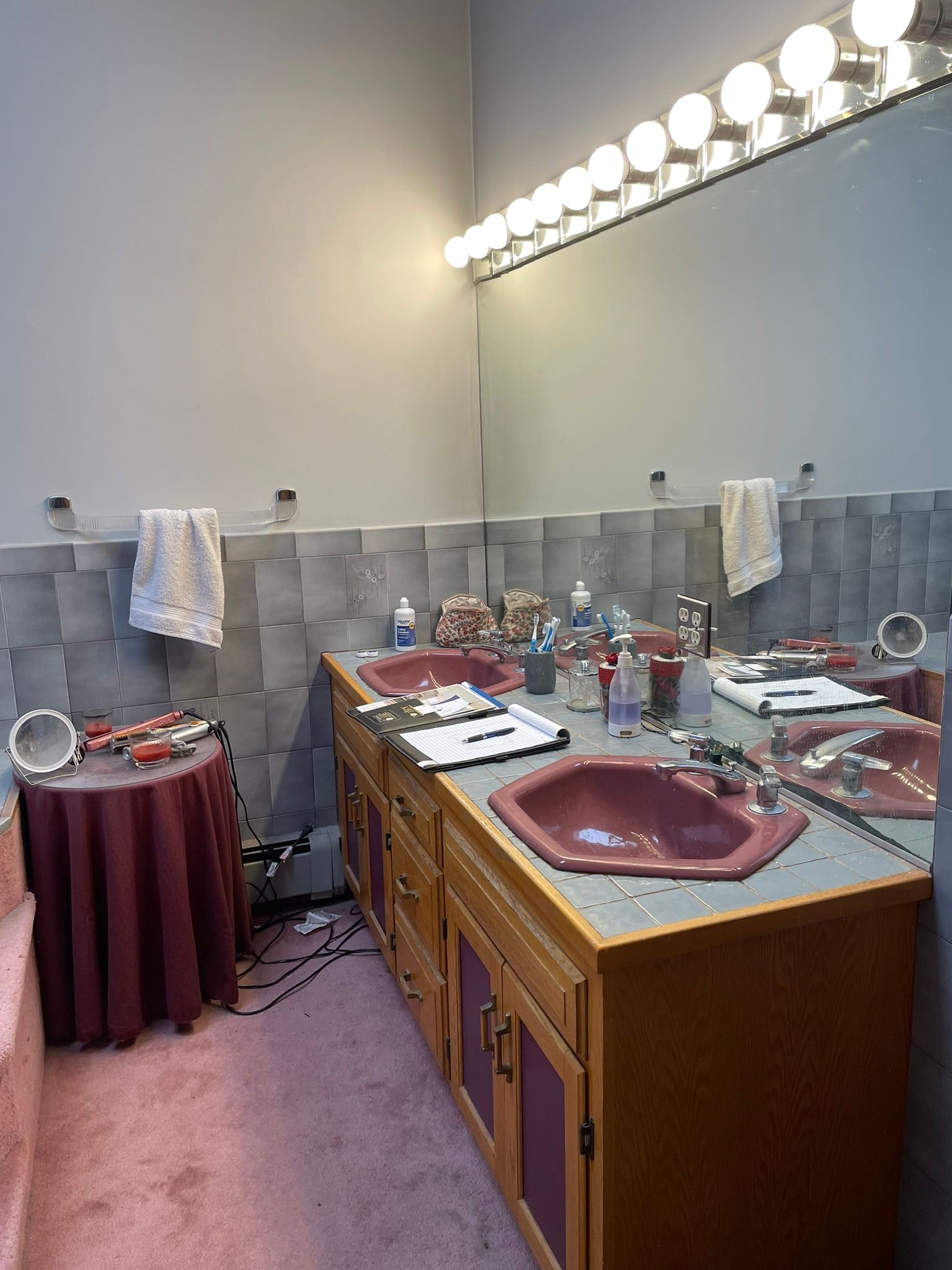 Bathroom vanity with mirror lights, two sinks, toiletries, and a pink floor mat