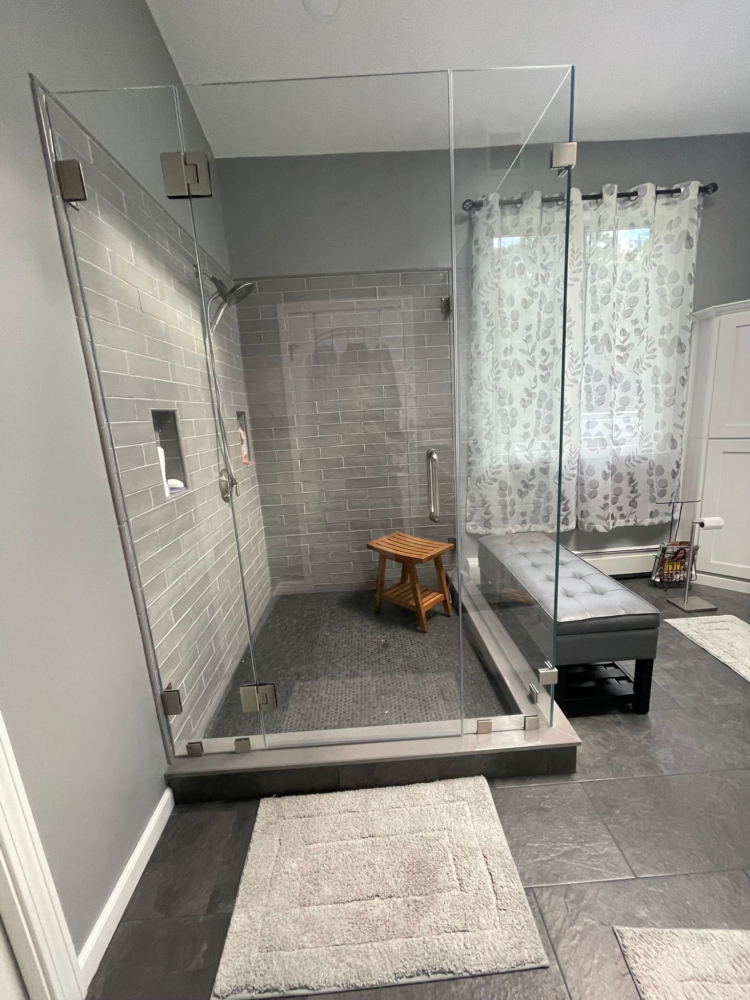 Glass-enclosed shower with gray tile walls and a small wooden stool in a bathroom