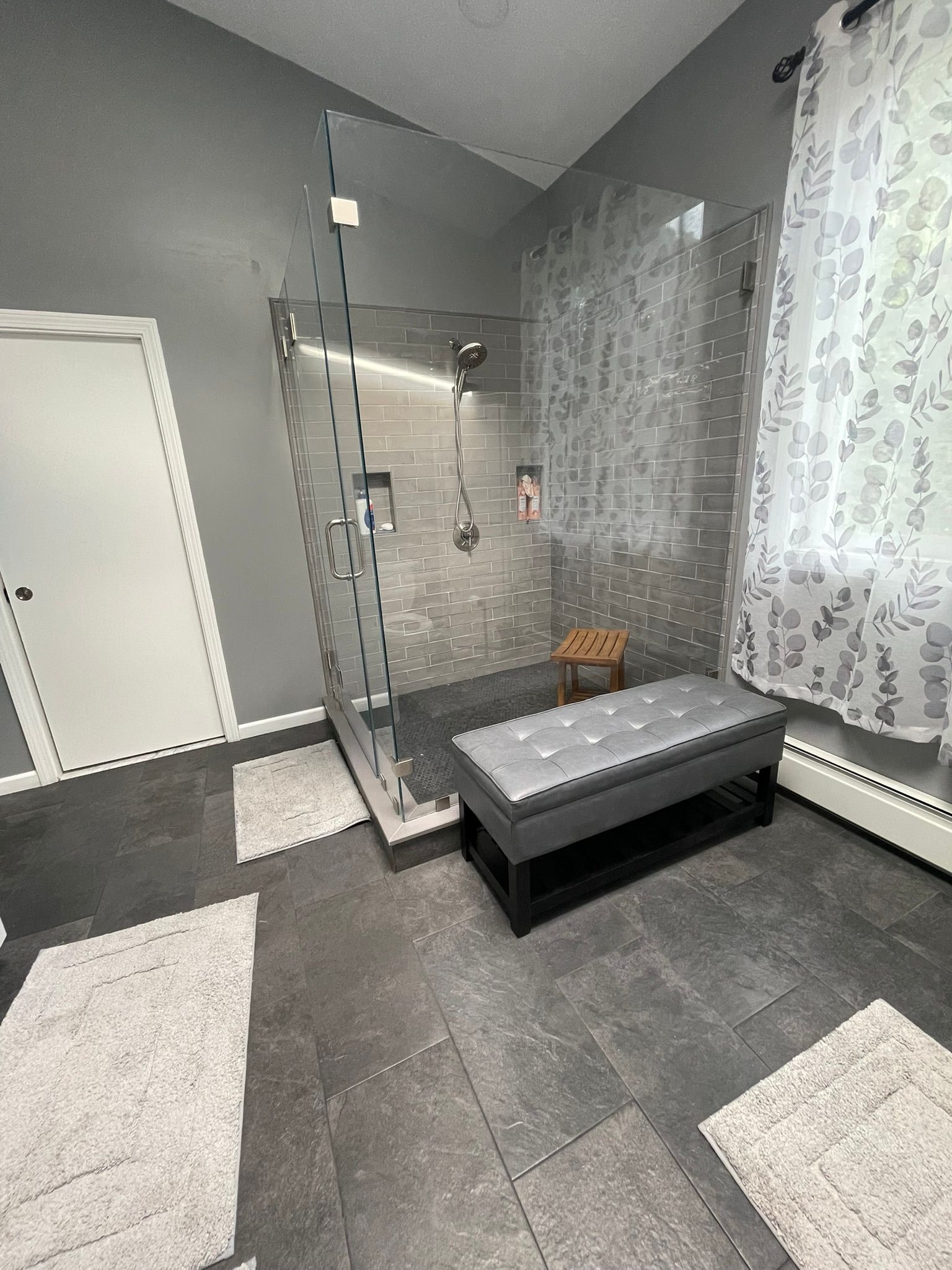 Modern bathroom with glass shower, gray tile, bench, and sheer curtains by a window.