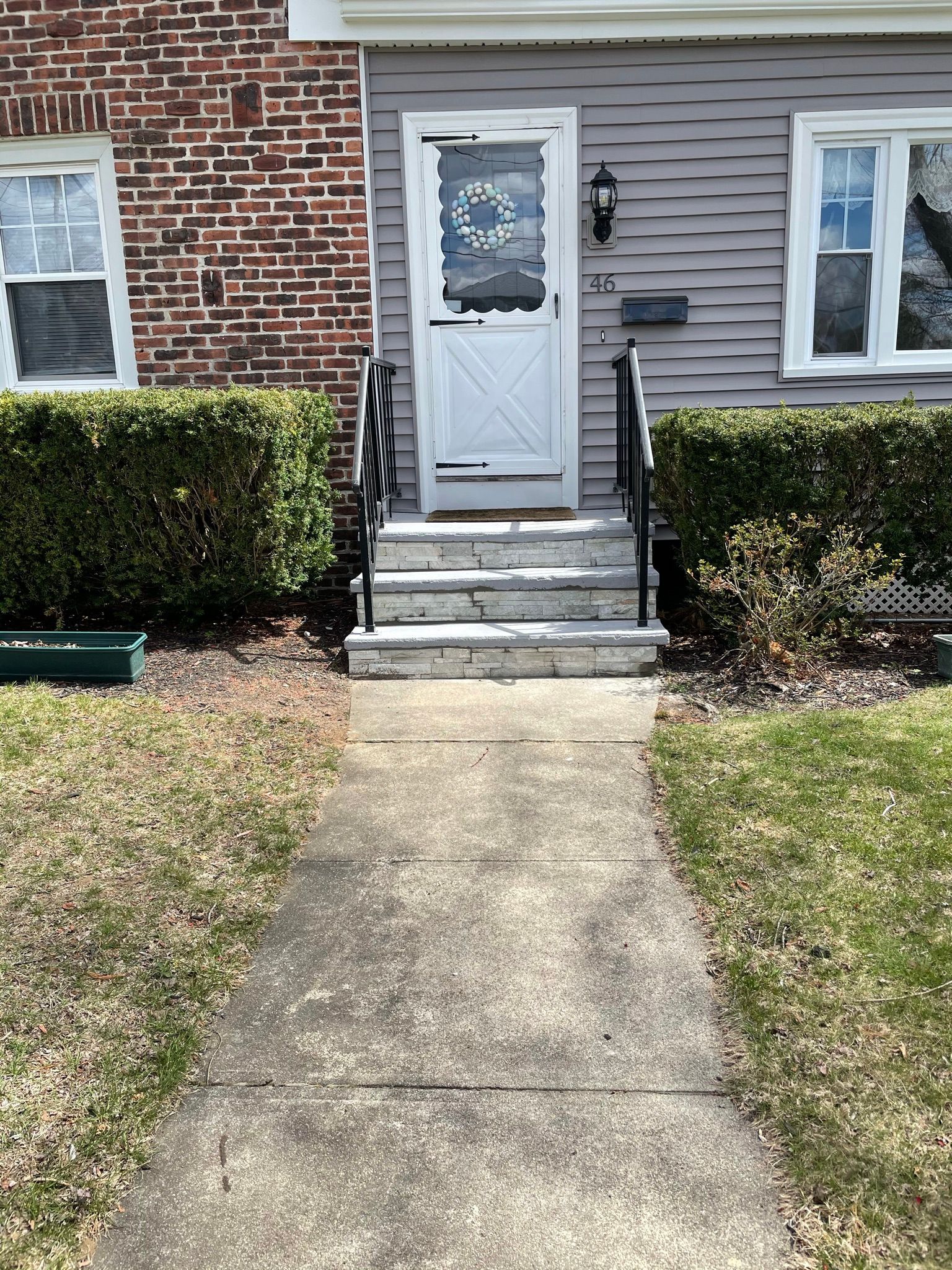 Front walkway leading to a brick house entrance with white door, steps, and trimmed hedges