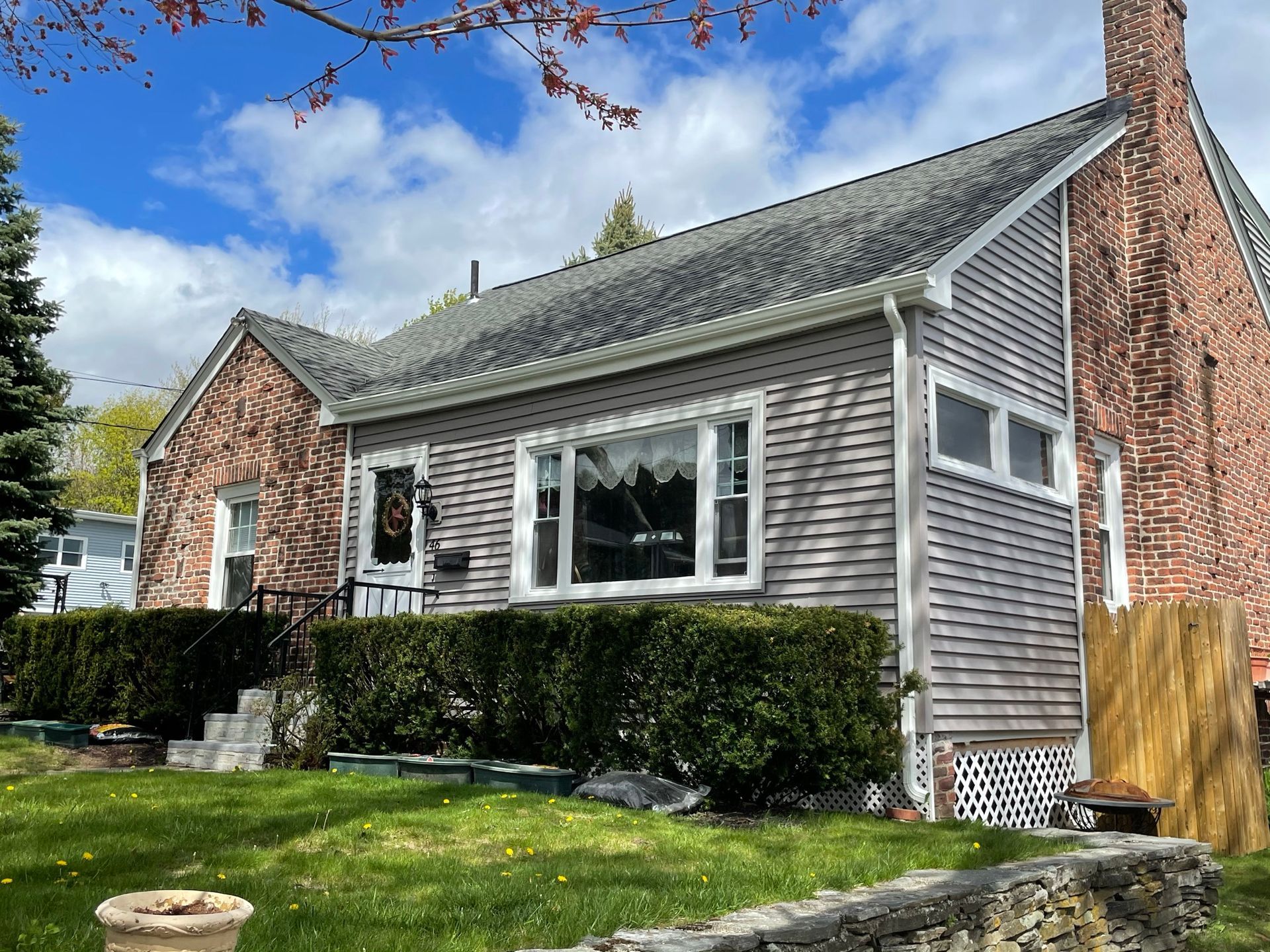 Gray shingle house with brick chimney, trimmed hedges, and lawn under a bright blue sky