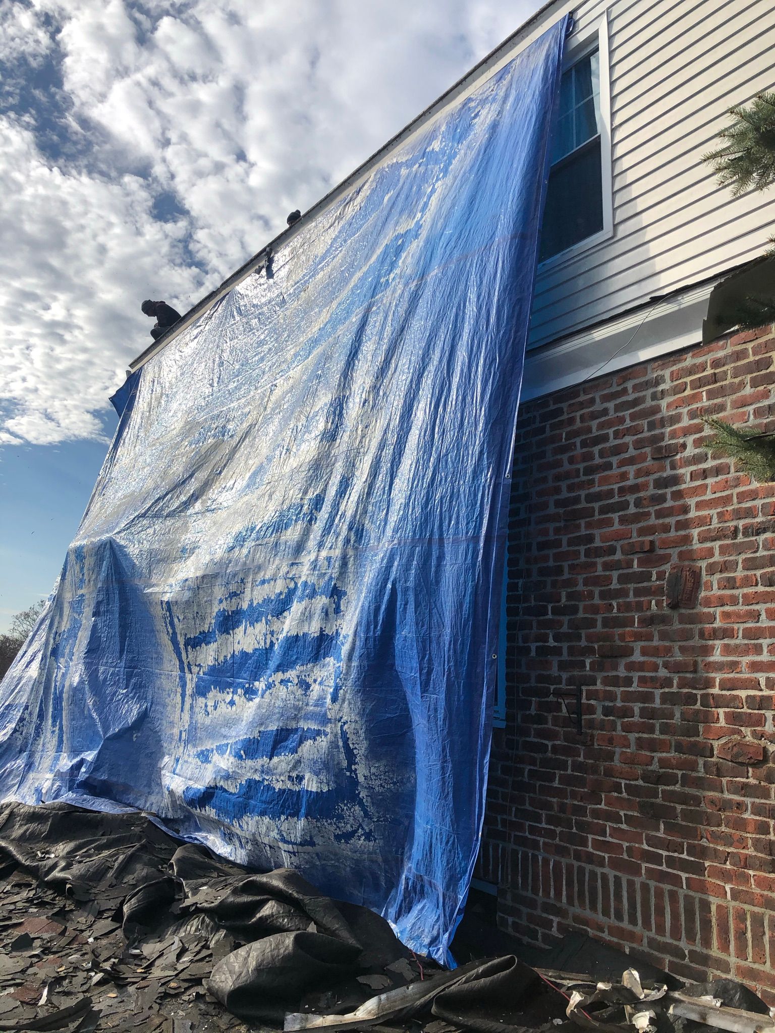 Blue tarp draped over a house exterior, covering damage beside a brick wall and debris pile.