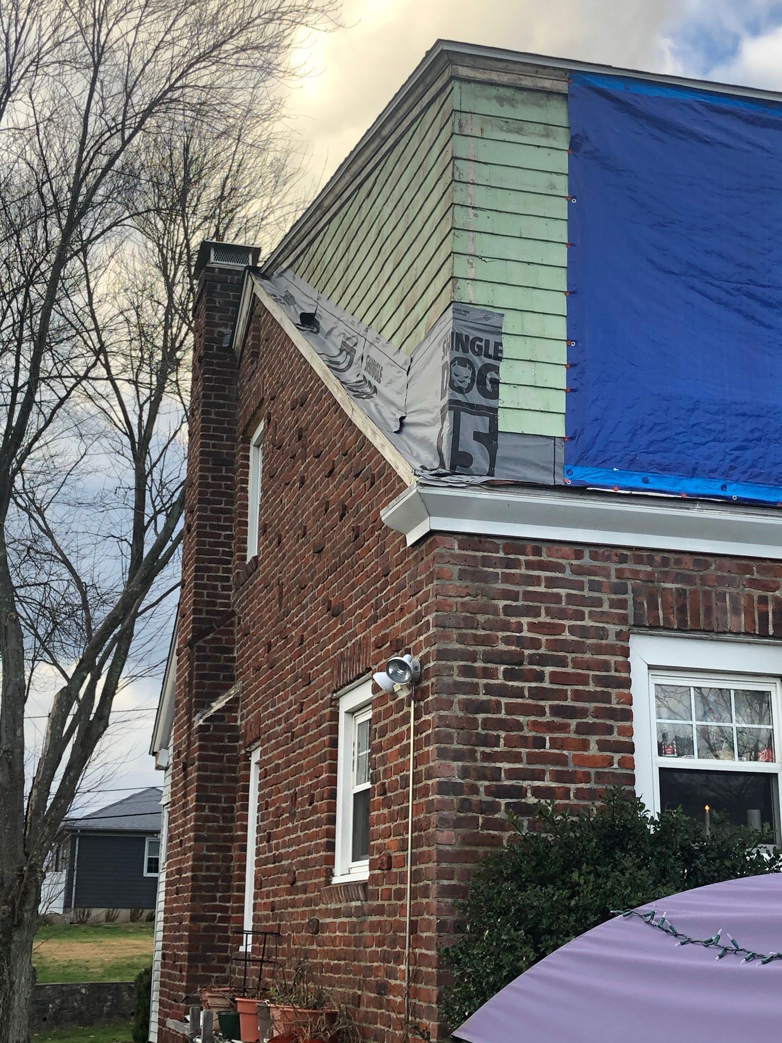 Brick house corner with a blue-tarp-covered roof and leafless trees against a cloudy sky