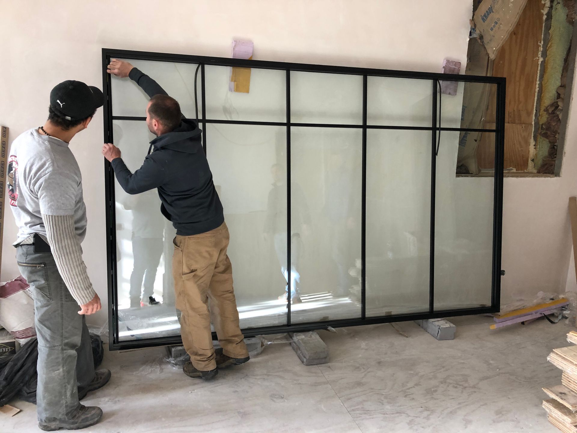 Two workers install a large black-framed glass partition in a room under renovation.