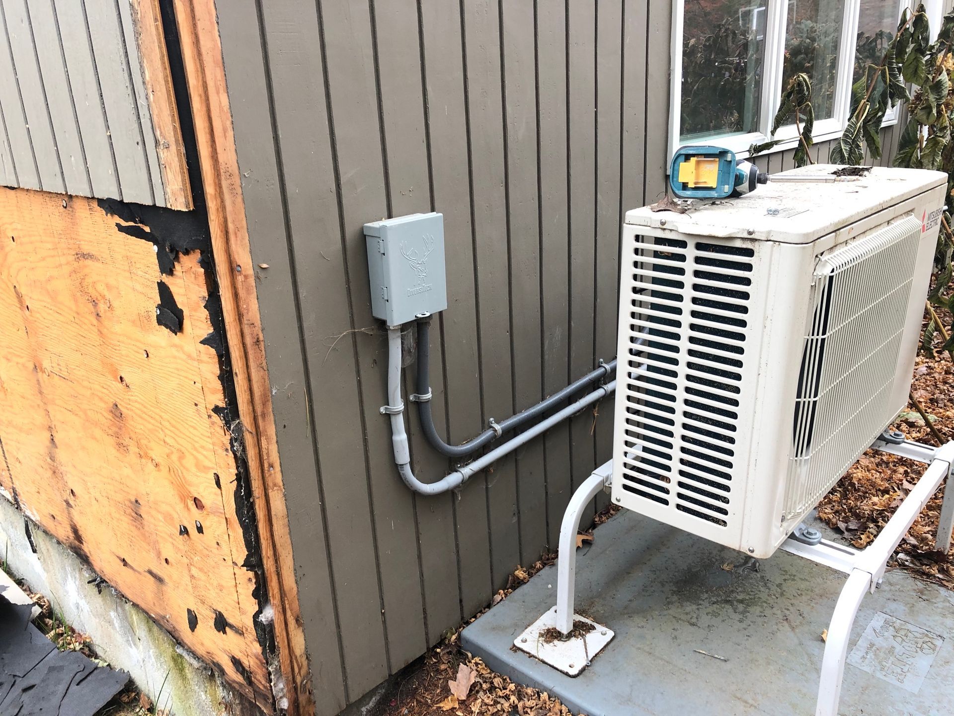 Outdoor HVAC unit beside a brown building wall with electrical conduit and damaged plywood panel