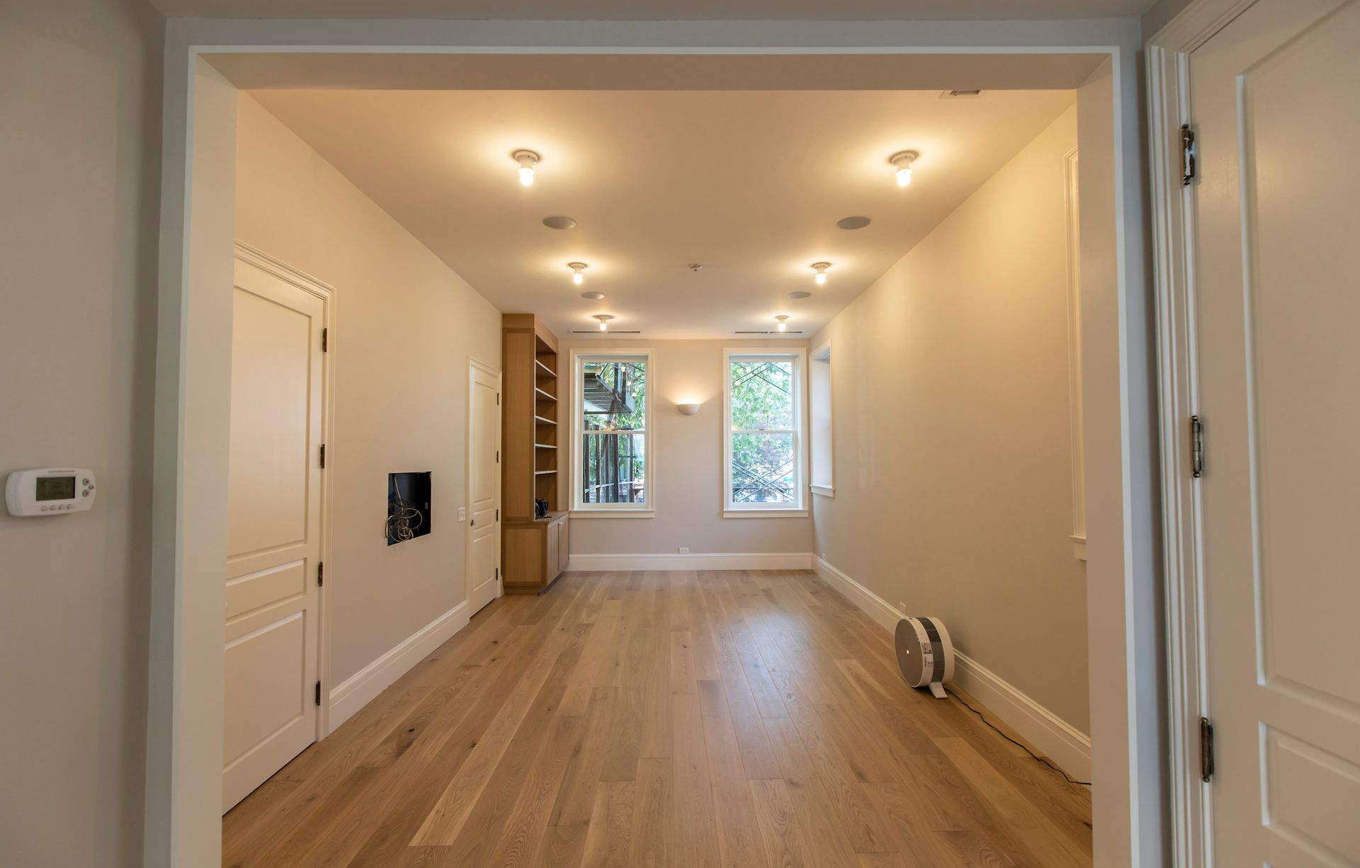 Empty renovated room with wood floors, recessed lights, and large windows at the far end