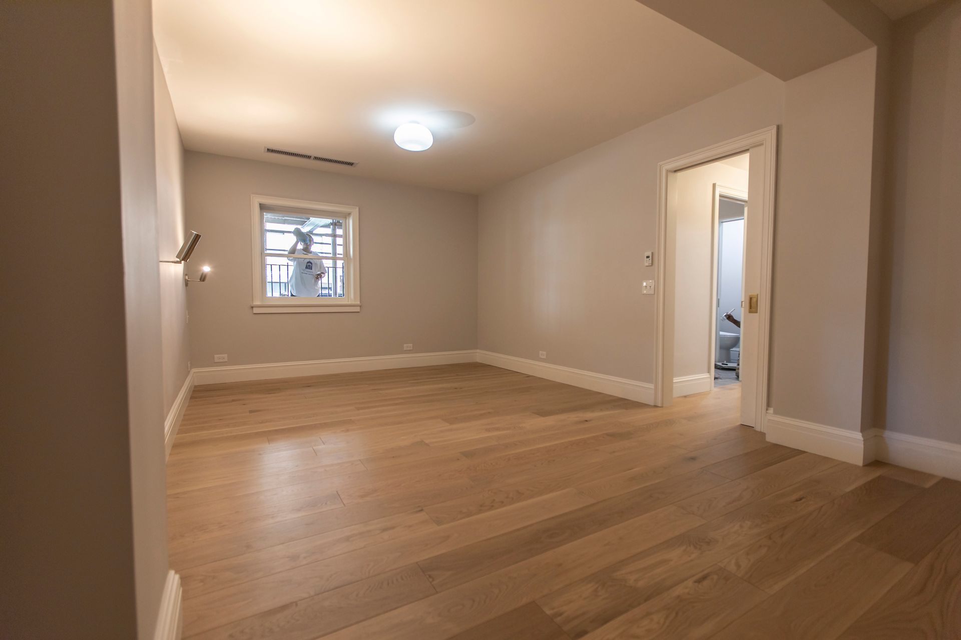 Empty beige room with hardwood floors, a ceiling light, and a small window.