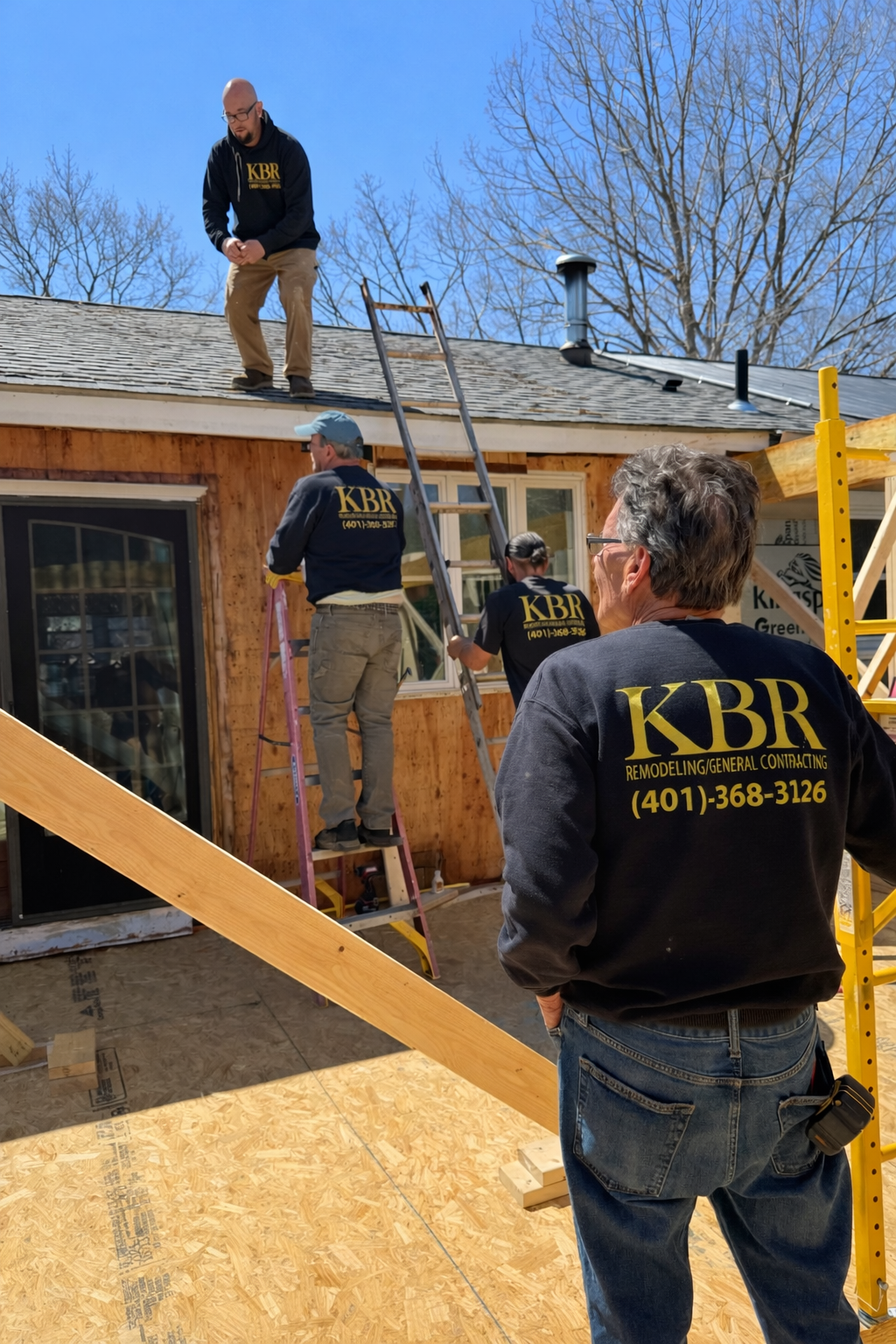 Construction workers on a roof and near a house; one man stands on the roof. Bright sunlight.