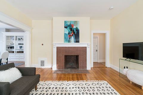 Living room with fireplace, abstract art, dark sofa, rug, wood floor, TV on glass table, white ottoman.