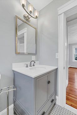 Gray bathroom vanity with white countertop, mirror, and light fixture. Open doorway leads to a hardwood floor.