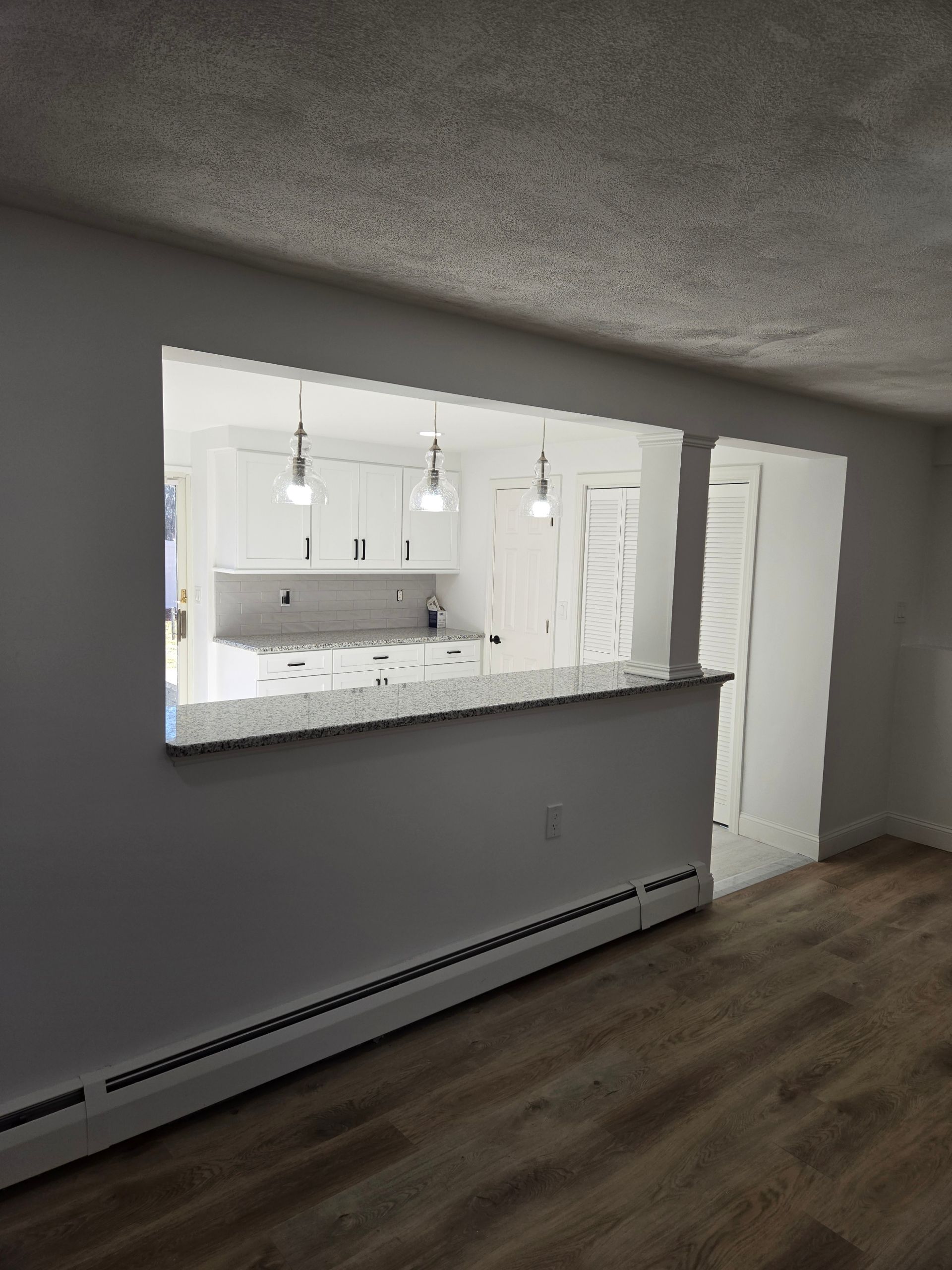 Interior view of a remodeled home with a pass-through to a white kitchen, featuring granite countertops and pendant lights.