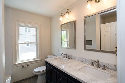 Bathroom with double sinks, marble countertop, silver framed mirrors, toilet, window, and black cabinets.