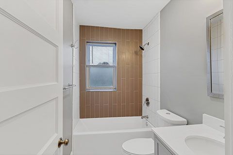 Bathroom with white and brown tiles, window, toilet, sink, and shower.