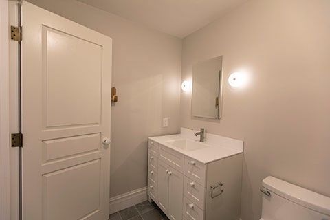 Bathroom with white vanity, door, mirror, and toilet; neutral walls and gray floor tiles.