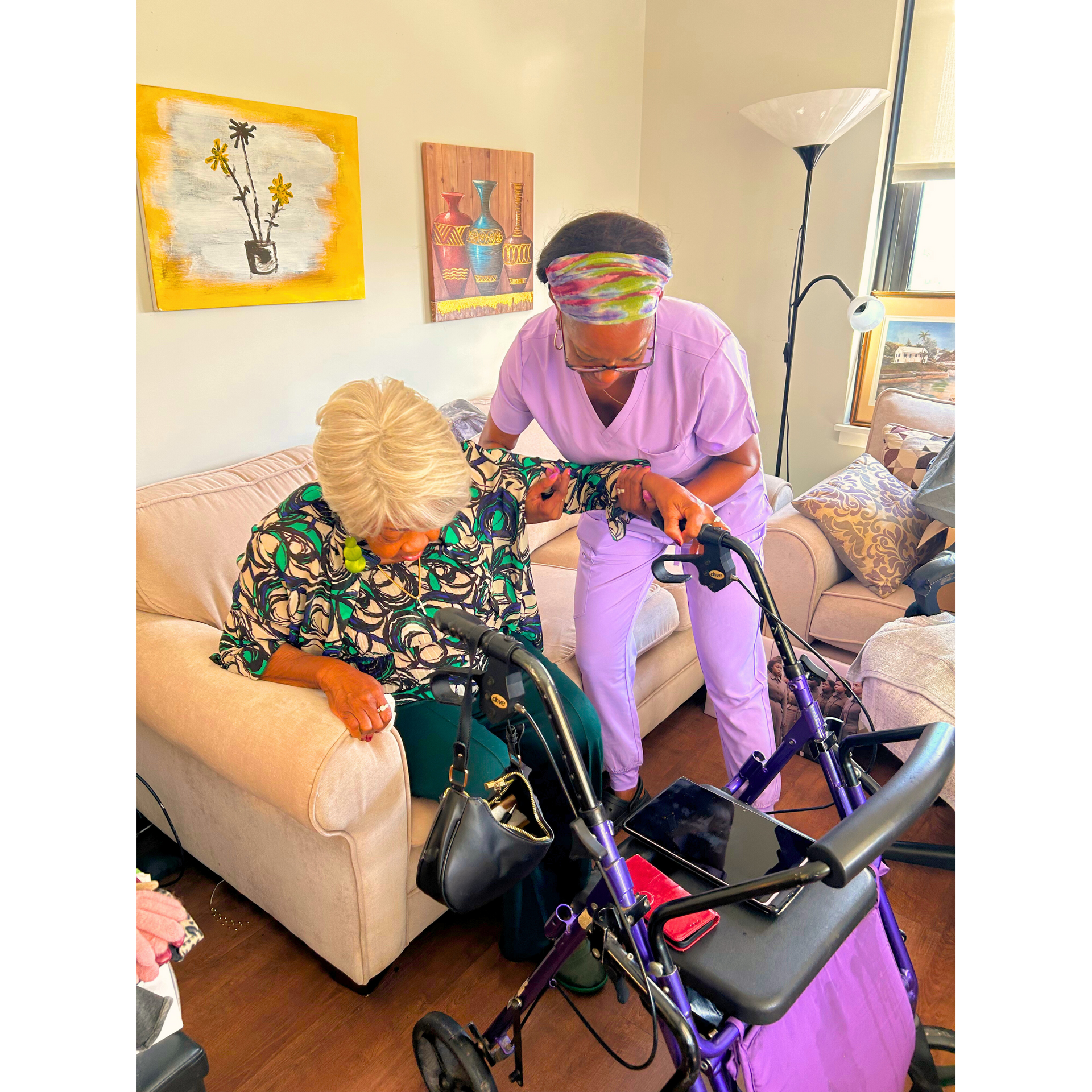 Two people in a living room with a walker and wheelchair, one seated on a sofa and one standing beside them.