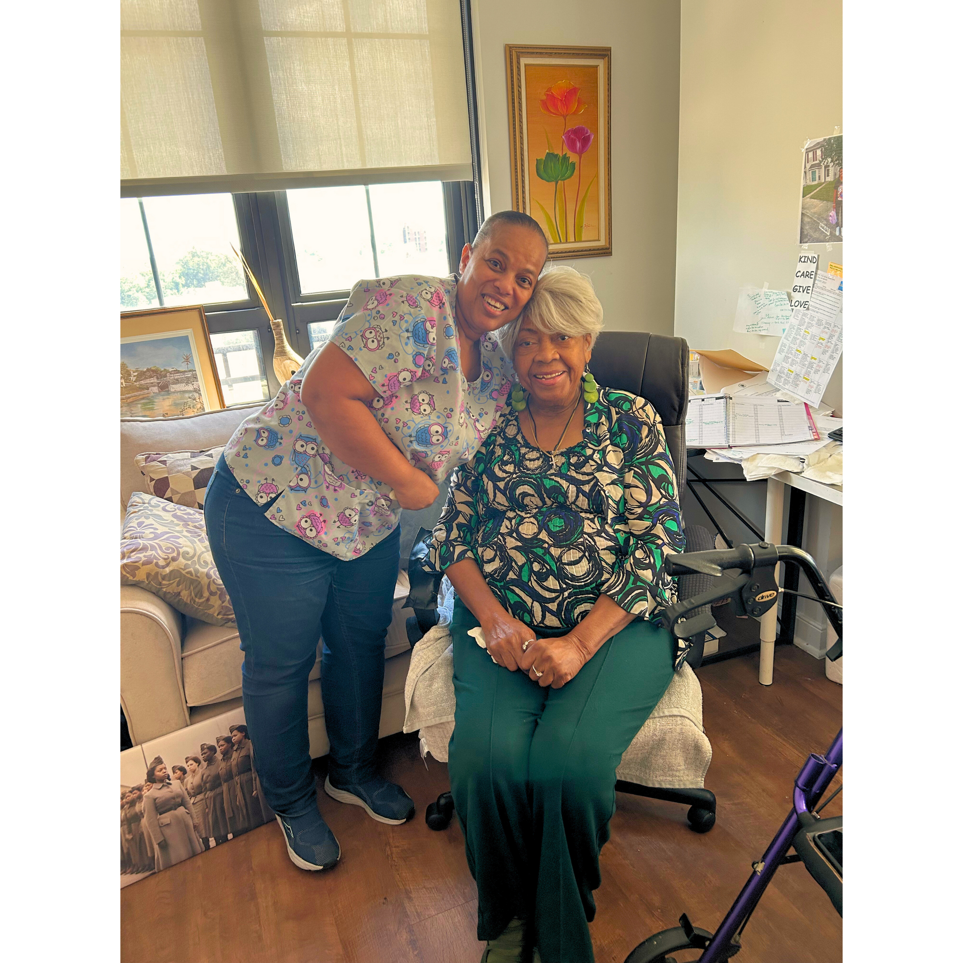 Two women smiling in a cozy office, one standing and hugging the other seated in a chair.