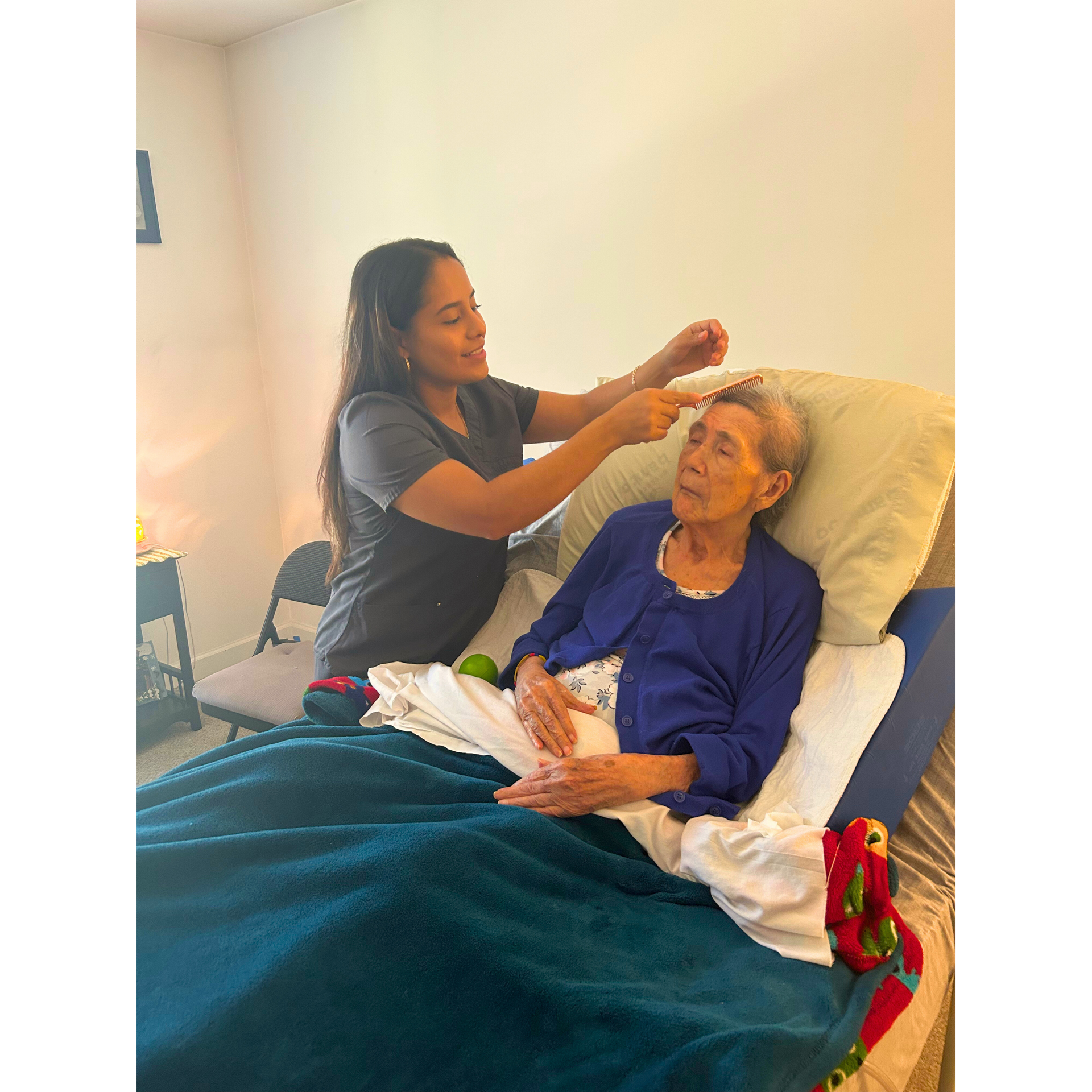 Caregiver adjusting an elderly patient's hair in a bed with white pillows and a teal blanket.