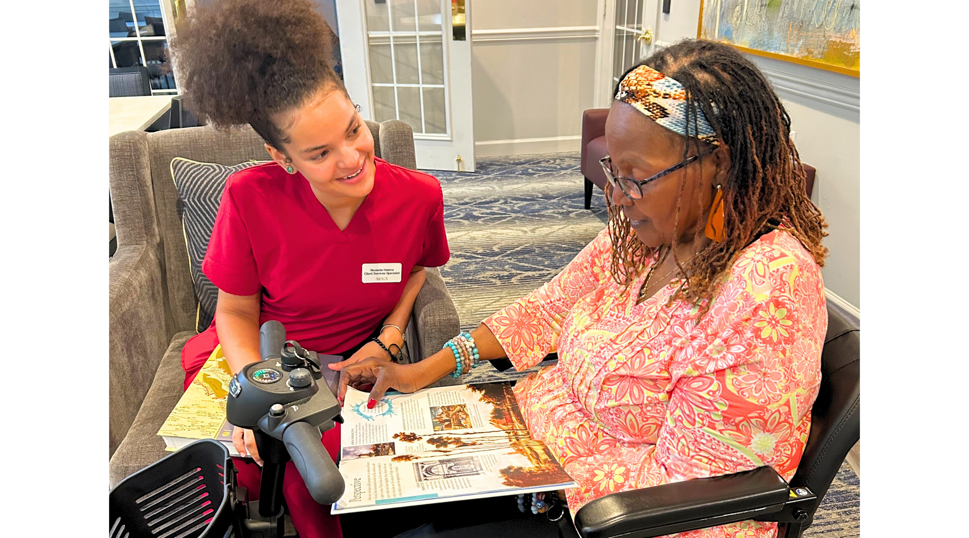 Woman in wheelchair reads with care worker in red scrubs.